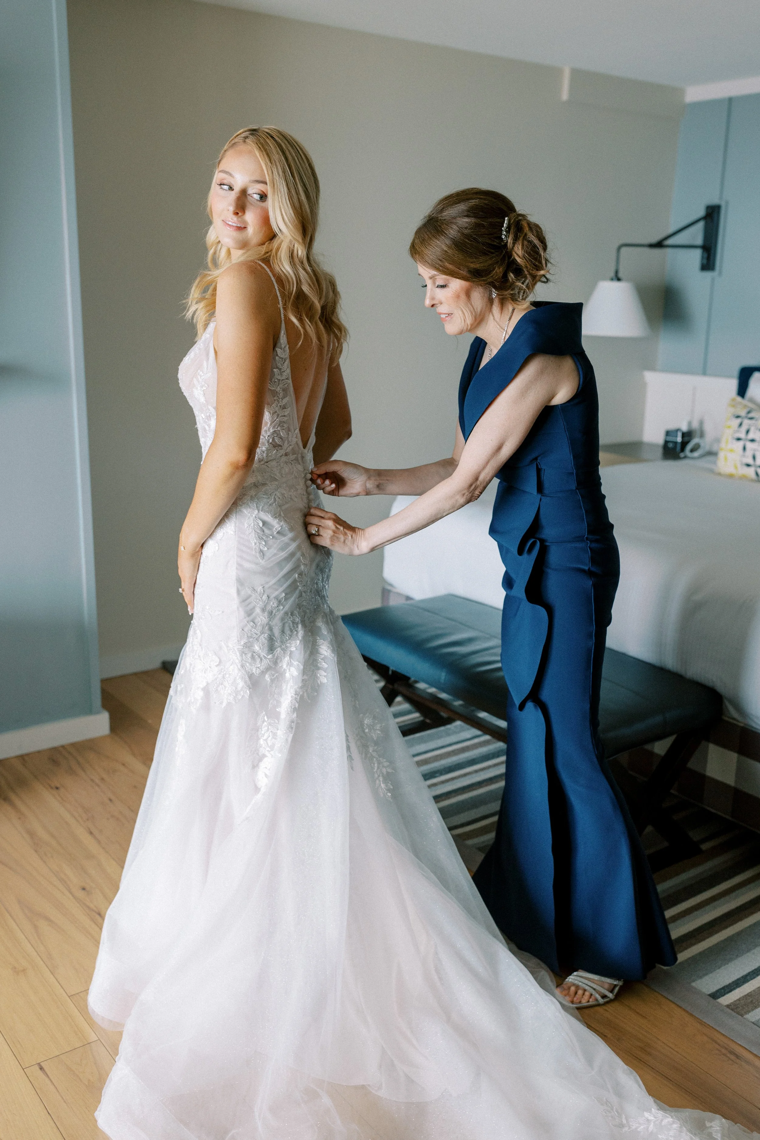 A bride in a lace wedding gown is being helped with her dress by a woman in a blue formal outfit inside a bedroom.