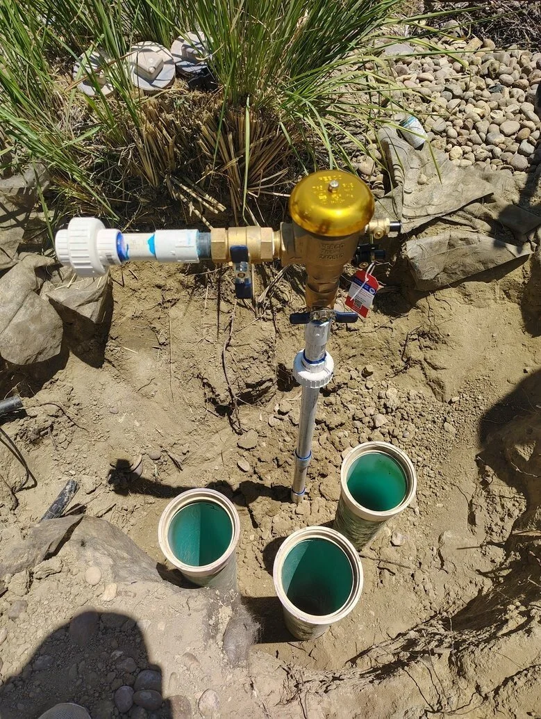 A water meter connected to a pipe system in the ground, with three pipes leading underground filled with green liquid, surrounded by rocks and dry soil.