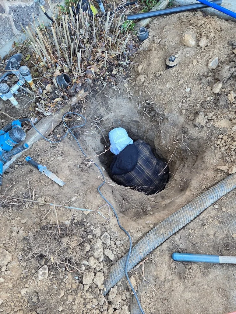 A person wearing a white hard hat and dark clothing working in a hole in the ground, with tools, pipes, and dirt surrounding the excavation site.