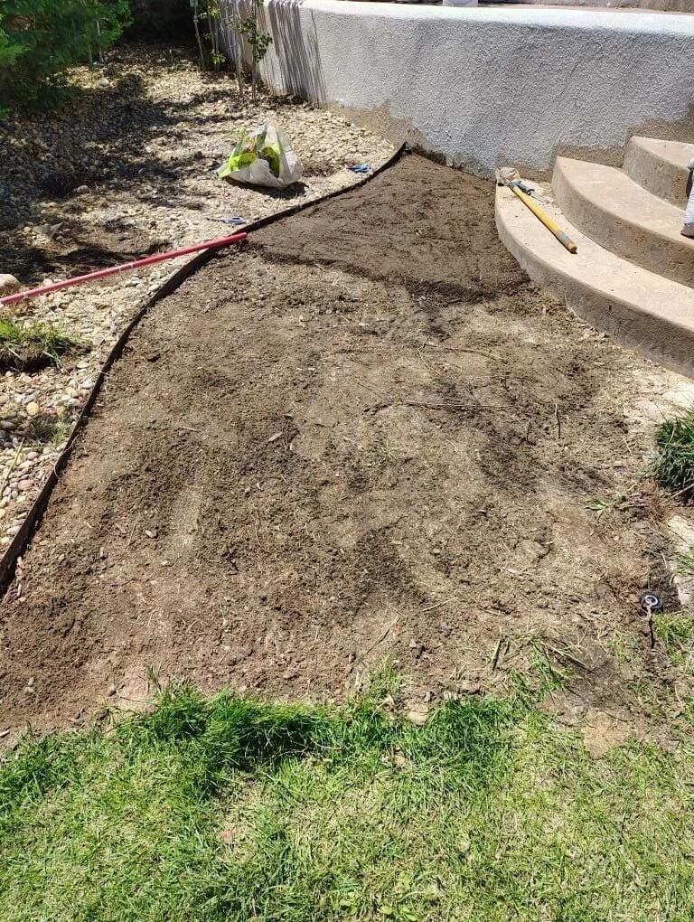 A backyard landscaping project with freshly tilled soil, a garden border, a level tool, and a garden hose, next to curved concrete steps leading to a house with a white wall.