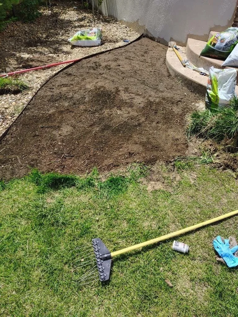 A backyard with a new landscaped area with freshly turned soil, a rake on the grass, and gardening supplies like bags of soil near the steps leading to the house.