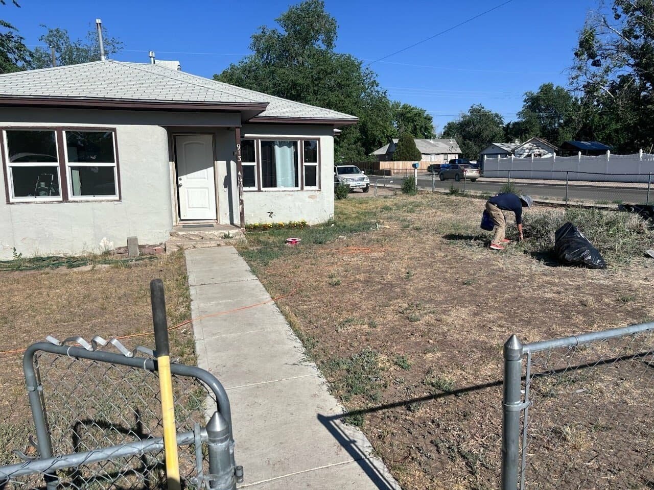 A person working in a barren front yard with trash bags, next to a small white house with three windows, a white door, and a pathway leading to the sidewalk, with a street and other houses in the background.