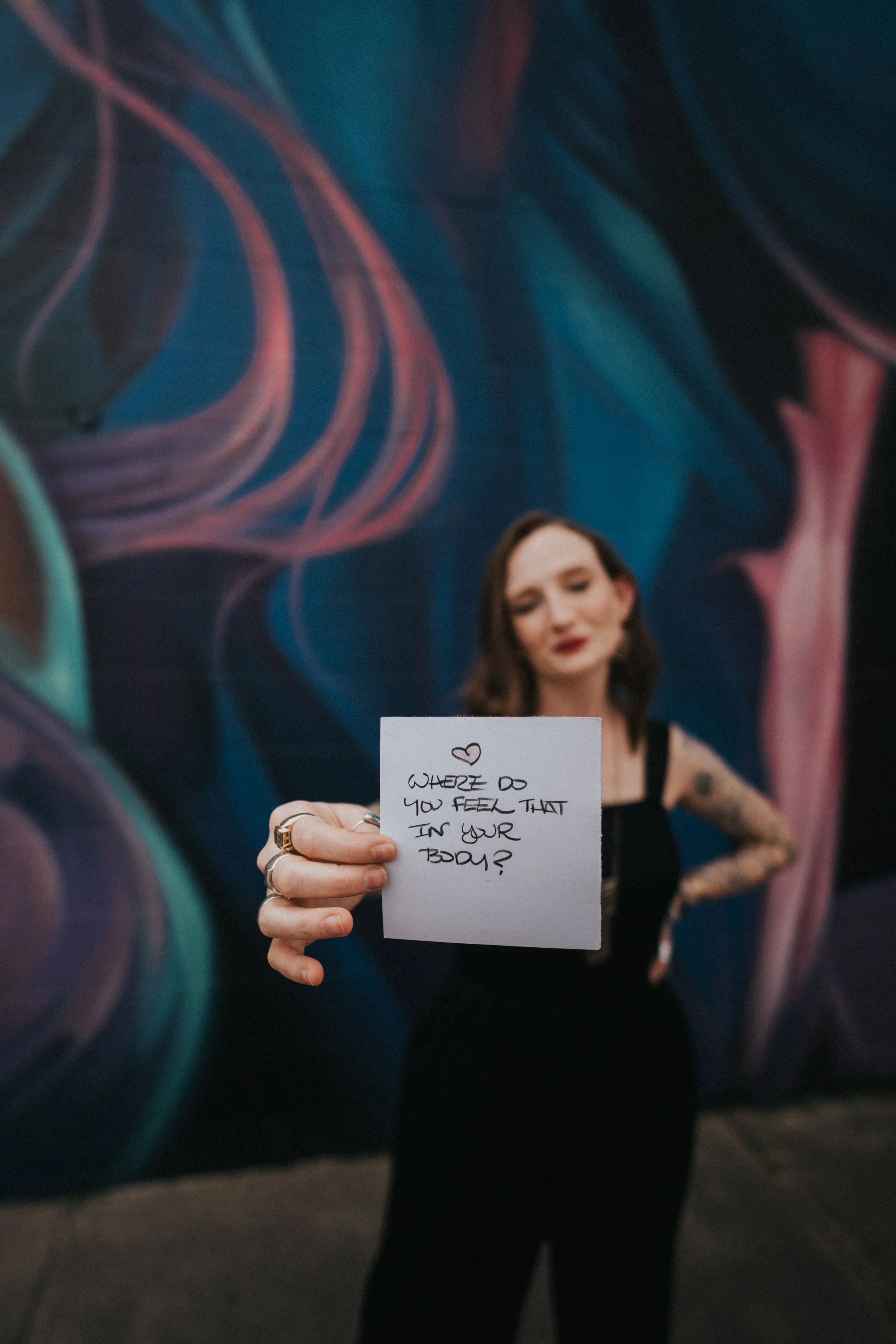 A woman with tattoos and dark hair standing against a colorful abstract mural, holding a note that asks, 'Where do you feel that in your body?'