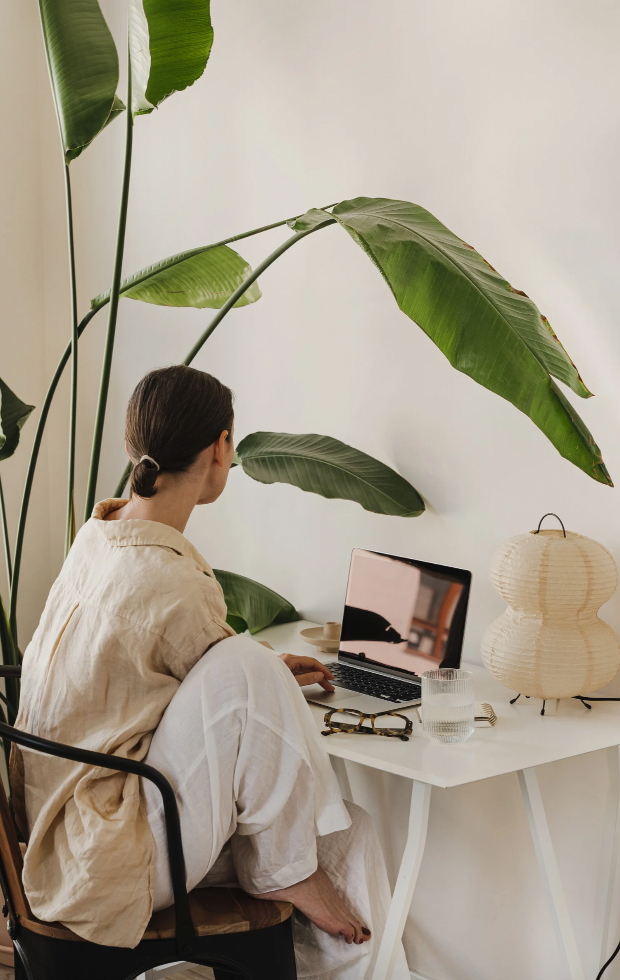 A woman sitting at a white desk with a laptop, glasses, a glass of water, and a paper lantern, surrounded by large green leaves of a potted plant. Telehealth private pay therapy services for Kentucky.