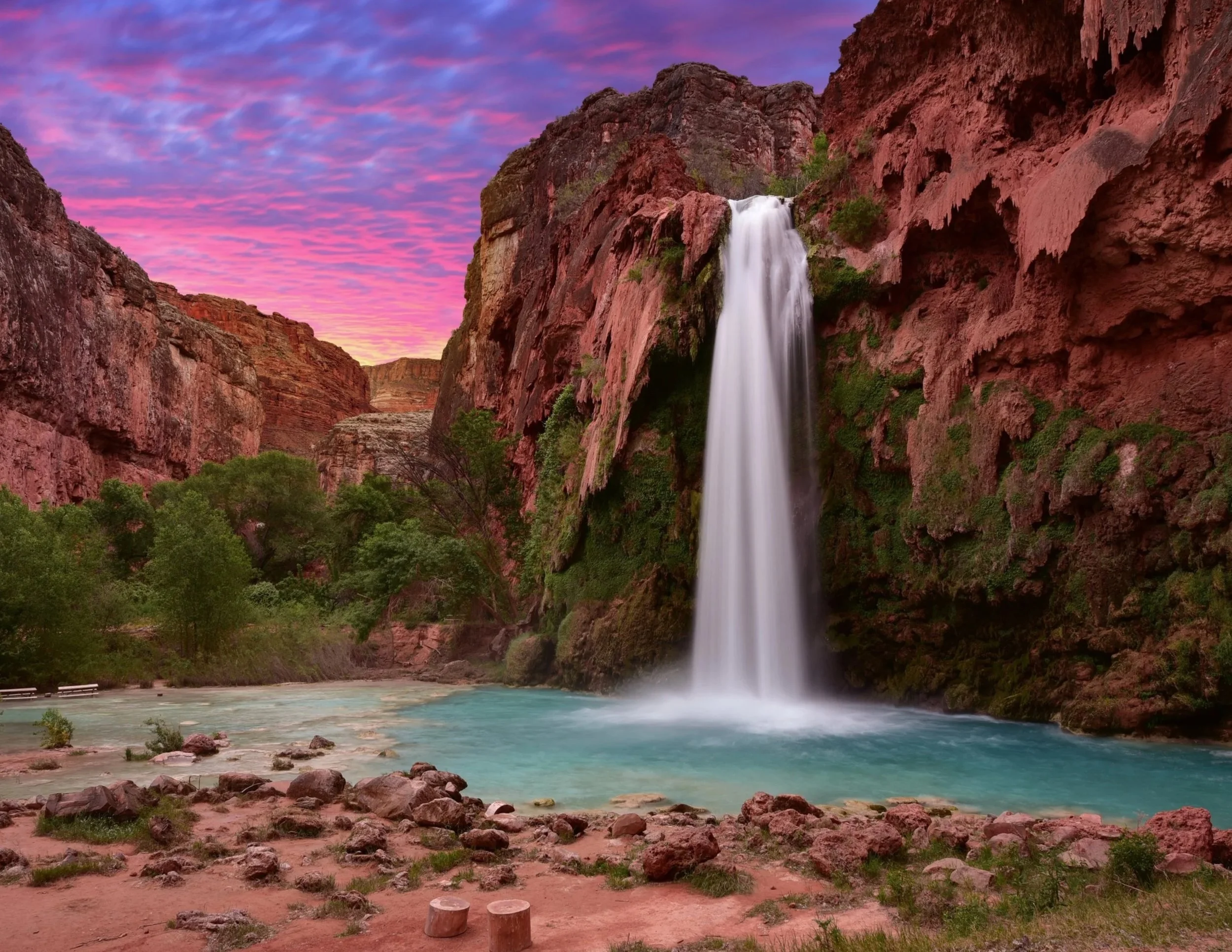 A tall waterfall cascading down red rocky cliffs into a turquoise pool below, with green trees along the shore and a pink and purple sky at sunset.