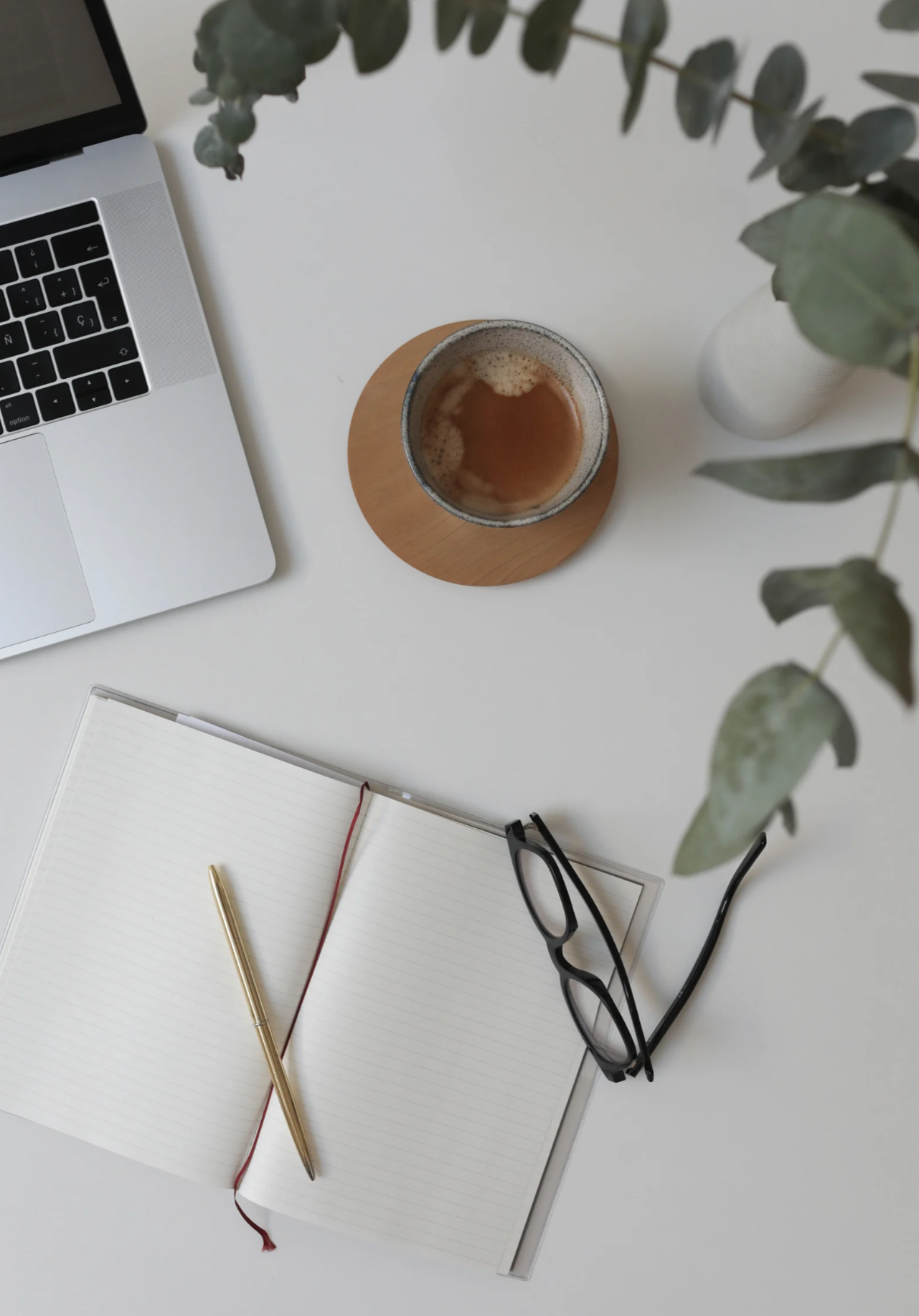 Top-down view of a white desk with a laptop, open notebook with a gold pen, a pair of black glasses, a cup of coffee on a wooden coaster, and a potted plant.