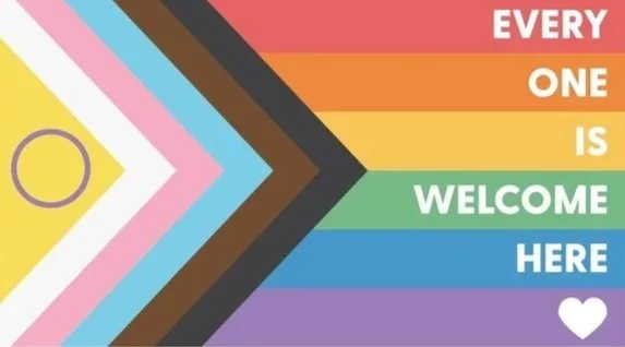 Colorful chevron pattern with rainbow-colored horizontal stripes and the text 'Every one is welcome here' with a white heart at the end. LGBTQIA+ safe space flag. All identities are welcome to do therapy with us.