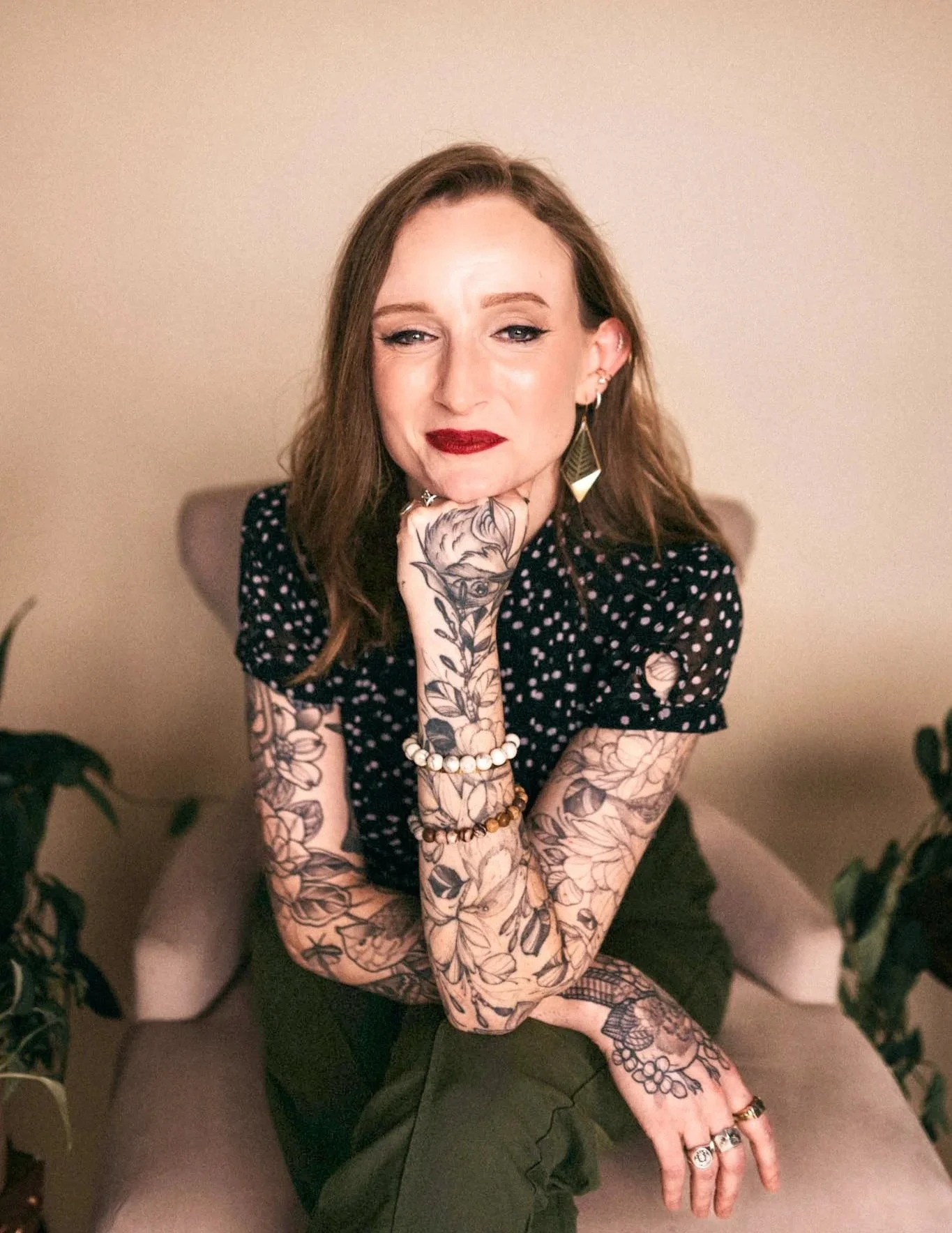 A woman with tattooed arms and hands, wearing a black polka dot top, sitting on a beige chair with her chin resting on her hand and smiling slightly, surrounded by plants. Chelsea McConaha, LCSW, who offers telehealth for Kentucky patients.
