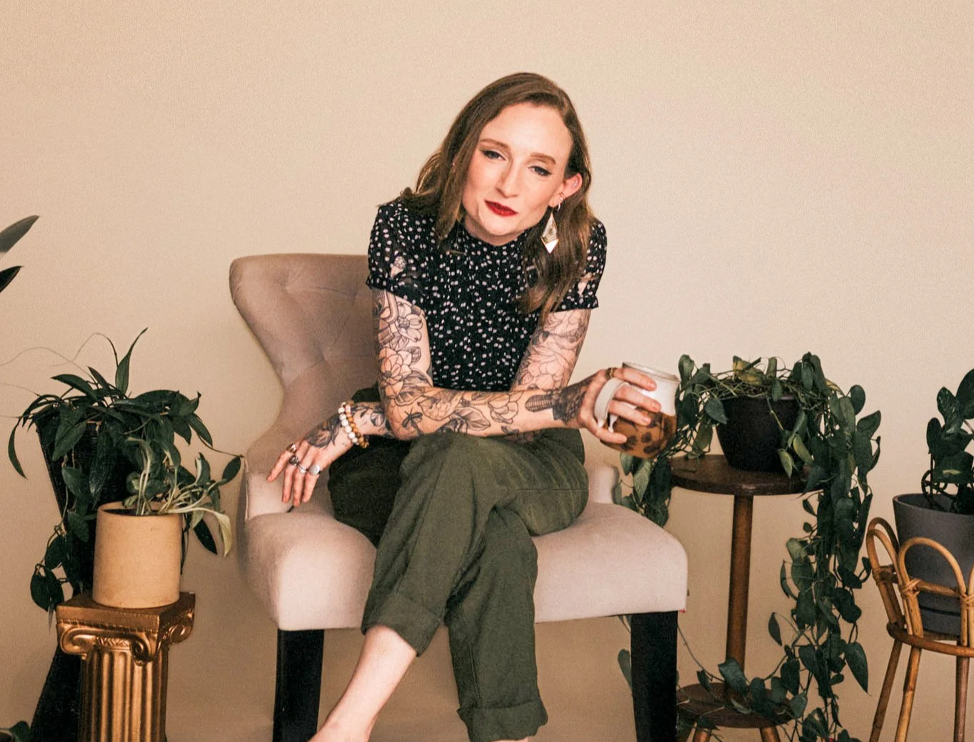 A woman sitting on a beige armchair with plants on either side, holding a cup, wearing a black polka dot blouse and green pants, with visible tattoos on her arms. Chelsea McConaha, LCSW, a mental health telehealth therapist in Kentucky.