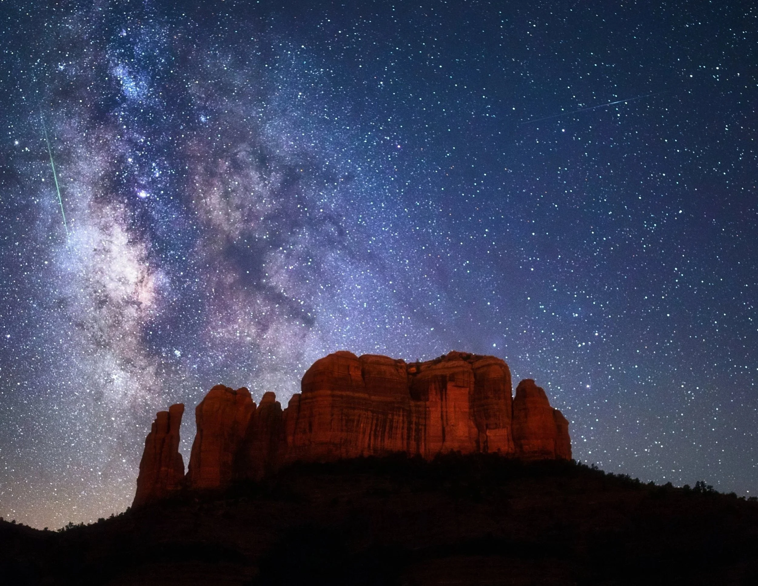 Night sky filled with stars and the Milky Way galaxy above a red rock formation in a desert landscape.