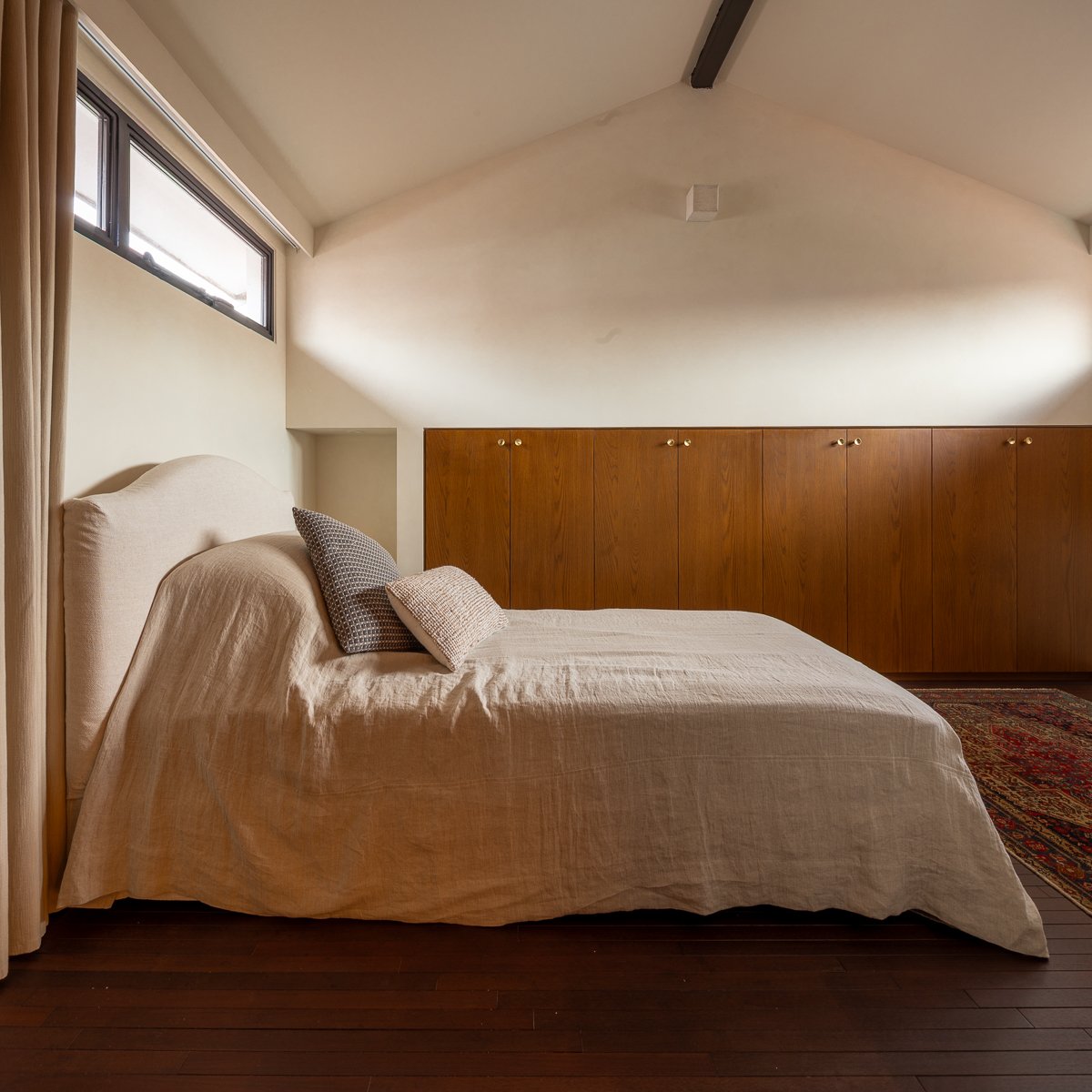 The room on the attic houses a single bed. Custom made low cabinetry are built on walls next to the bed. Similar dark toned wood flooring is used.