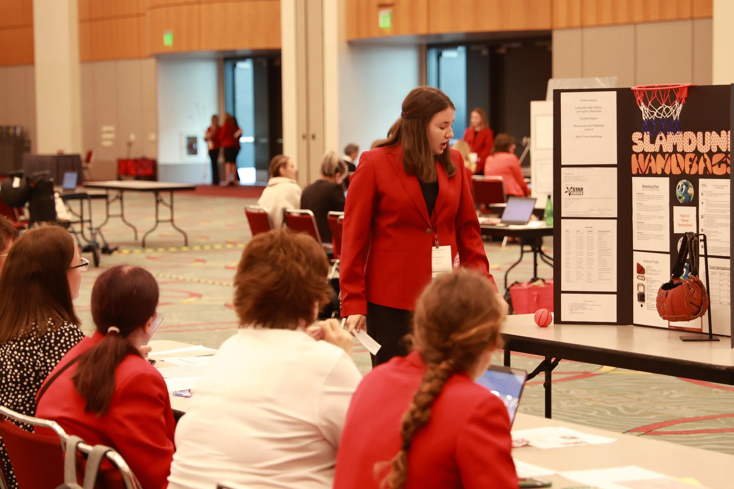 Women in red jackets attending a conference, sitting and talking around tables with presentation boards in a large conference hall.