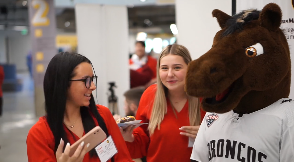 Three women and a person in a horse mascot costume are at an indoor event. The women are smiling and holding plates with food, one is looking at her phone. The woman in the center has long blonde hair and is wearing a red shirt. The woman on the left has dark hair, glasses, and is wearing a red shirt. The horse mascot is wearing a white sports jersey with 'Broncos' and an NFL logo.