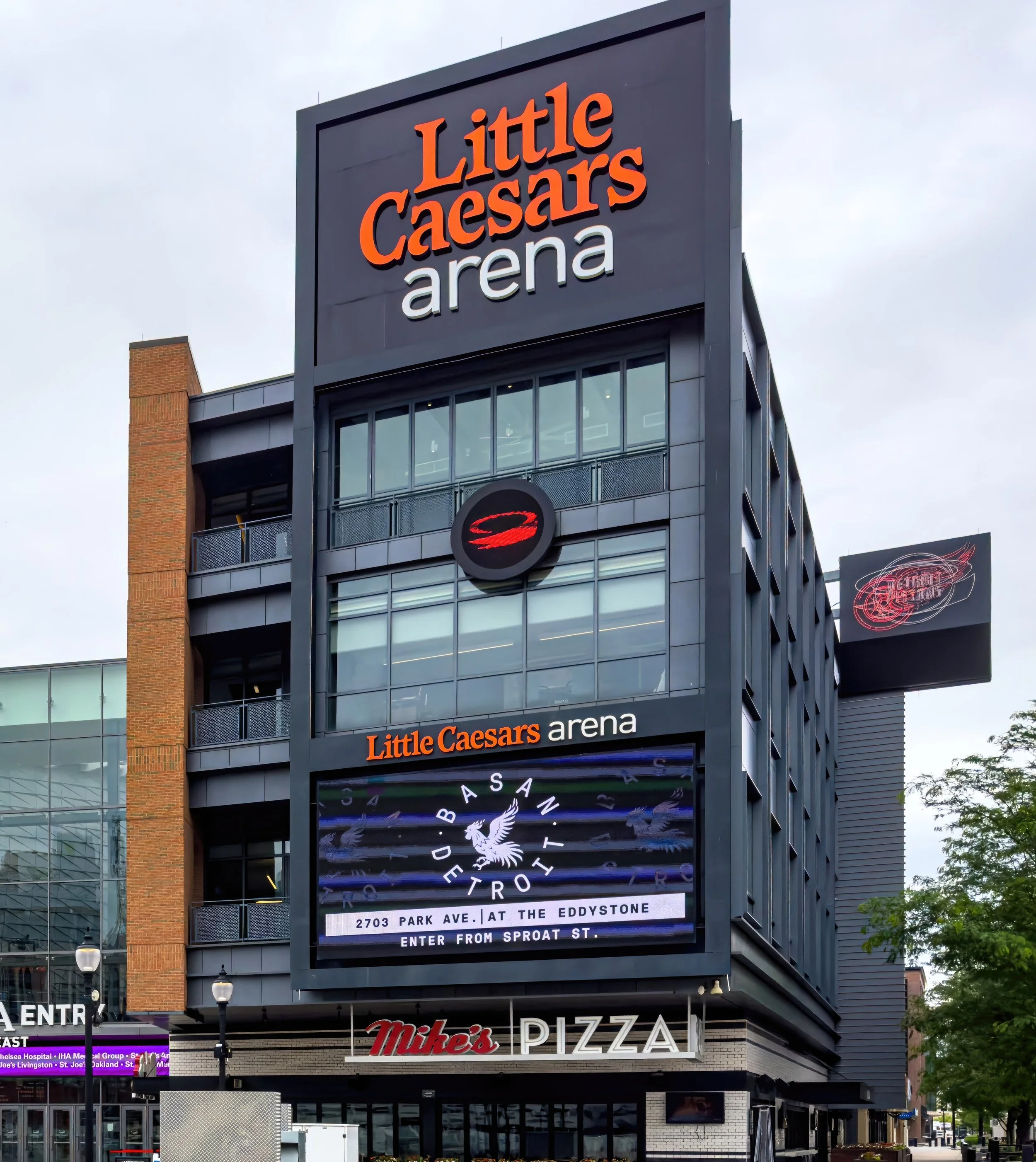 Exterior of Little Caesars Arena with digital sign displaying information about a Detroit basketball game and nearby Mike's Pizza restaurant.