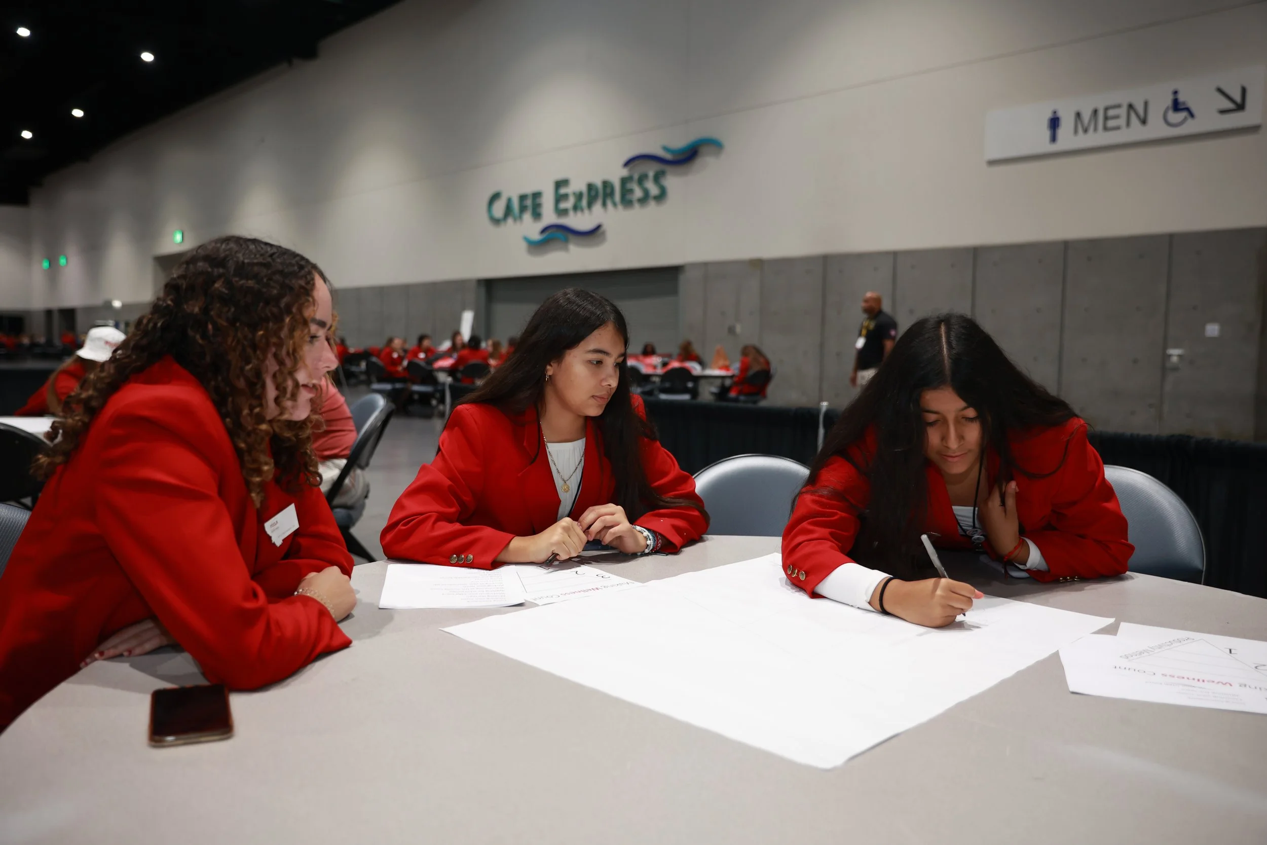 Three women dressed in red jackets sitting at a table, working on papers, in a large indoor convention center or conference space with a sign reading 'Café Express' in the background.