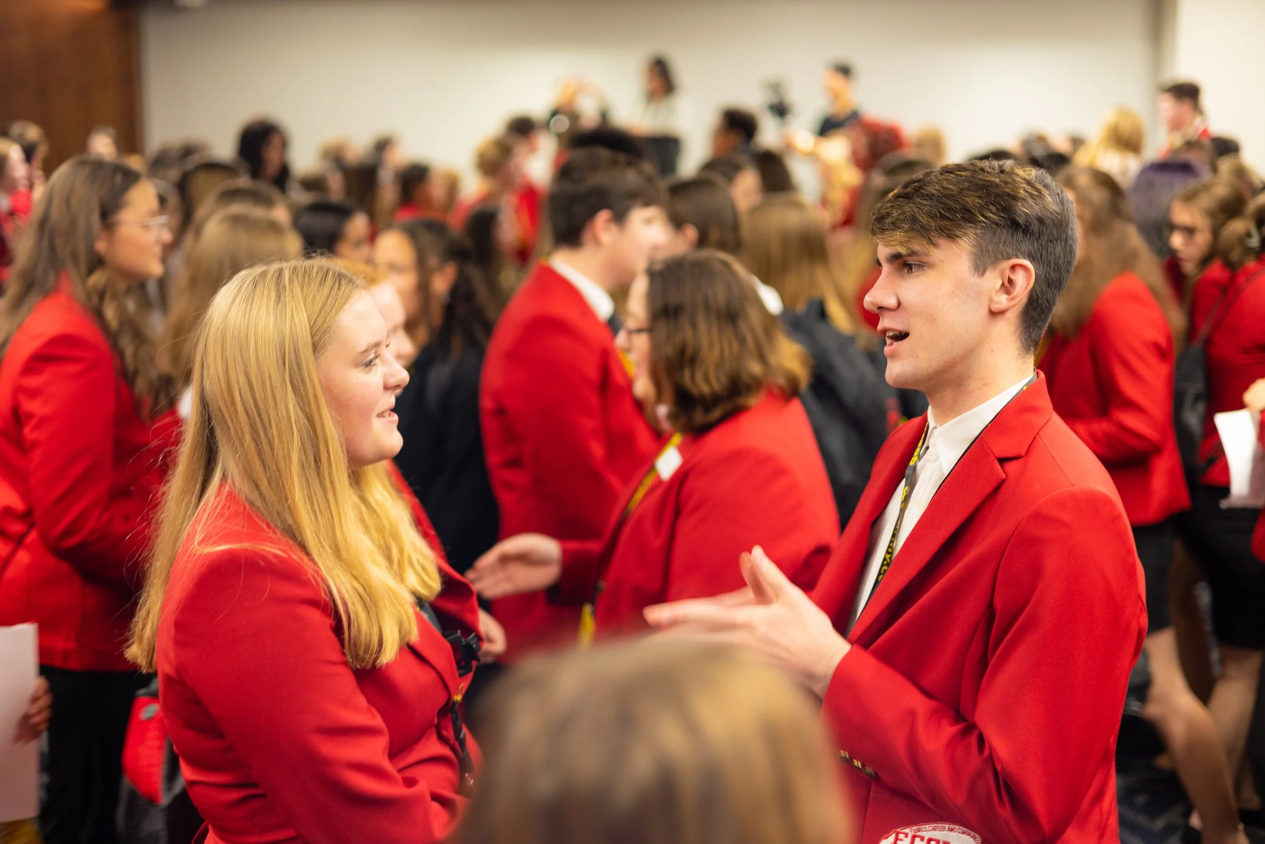 Students wearing red blazers at an indoor event, engaging in conversations and shaking hands.