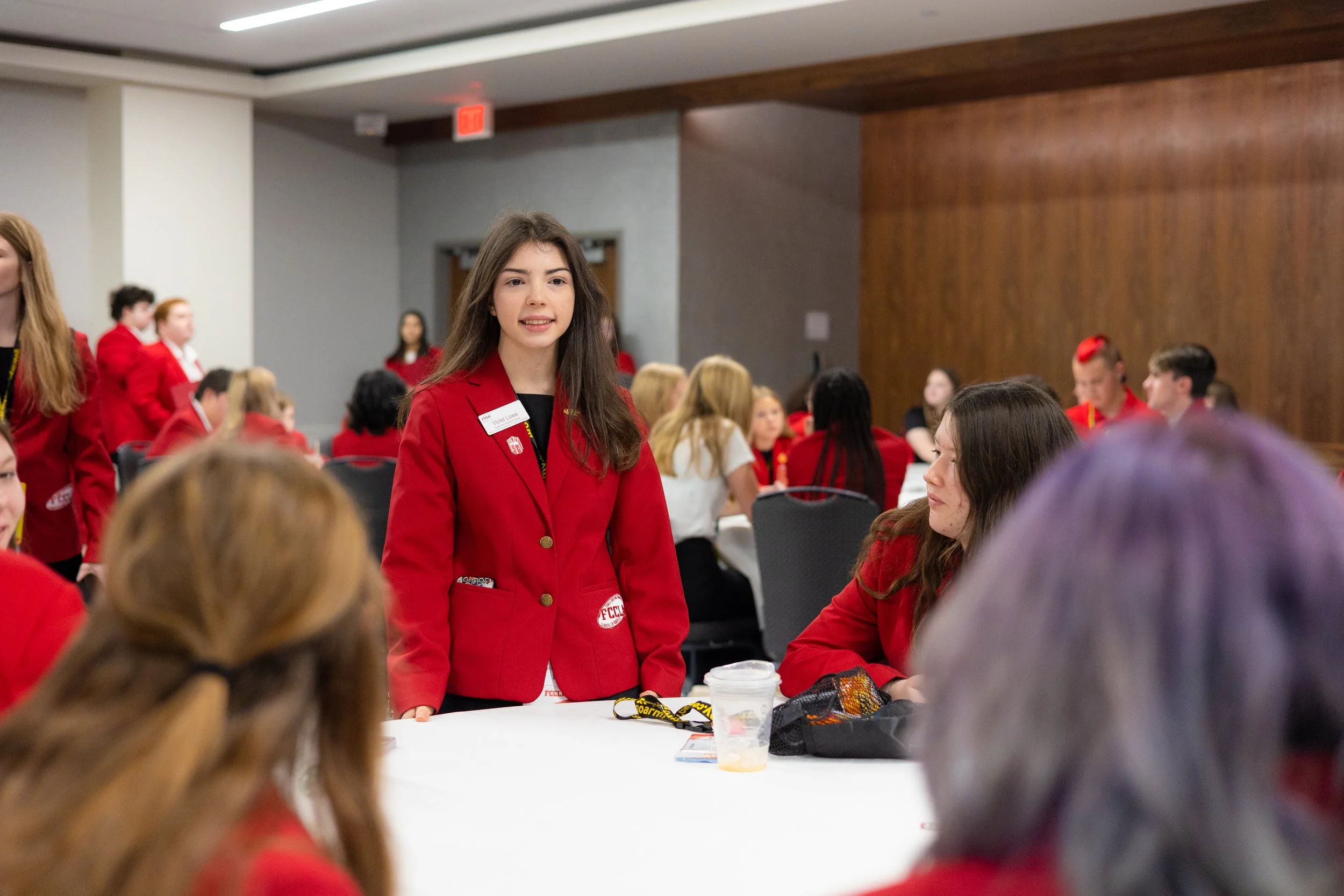 Young woman in a red blazer standing at a table with others seated around, all wearing red blazers, in a large room with wood paneling and gray walls, at a gathering or conference.