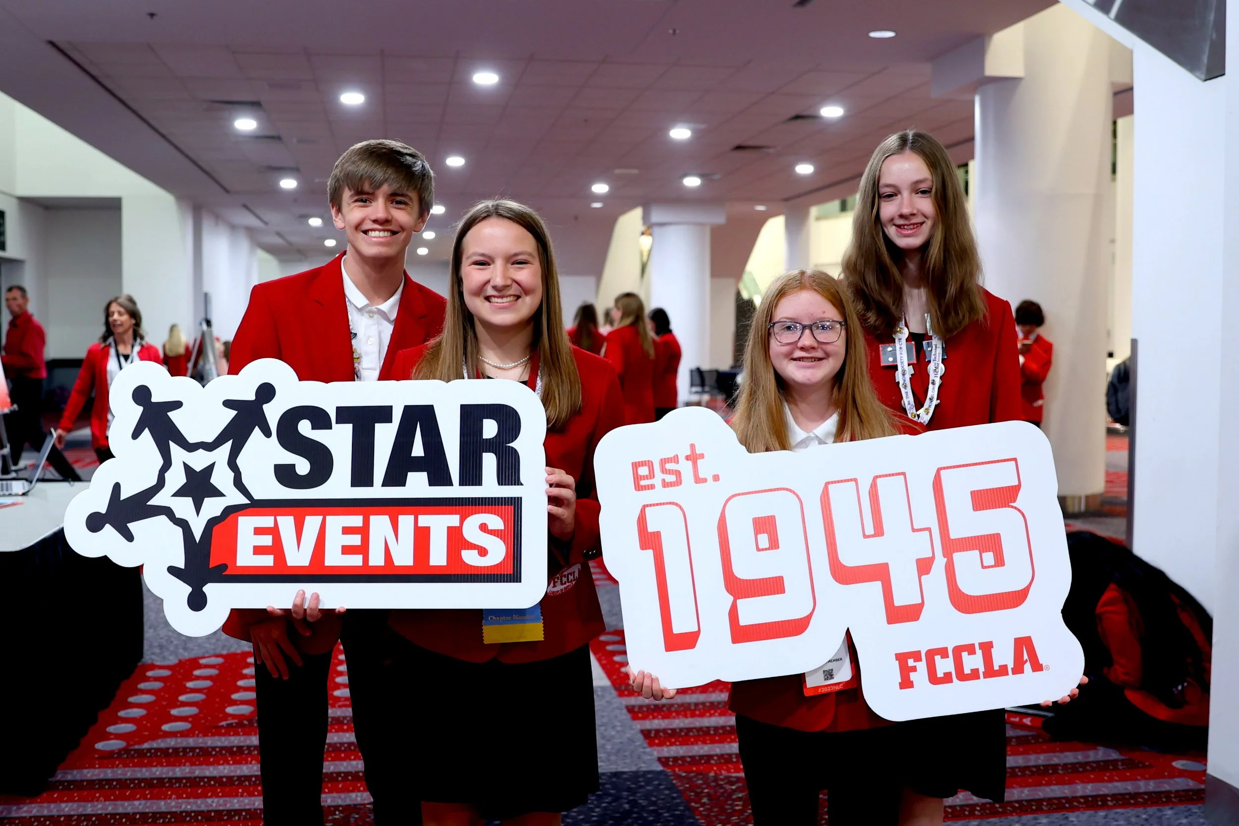 Group of four smiling young girls wearing red jackets at an indoor event, holding signs promoting the FCCLA organization and event. Background shows other participants and a decorated venue.
