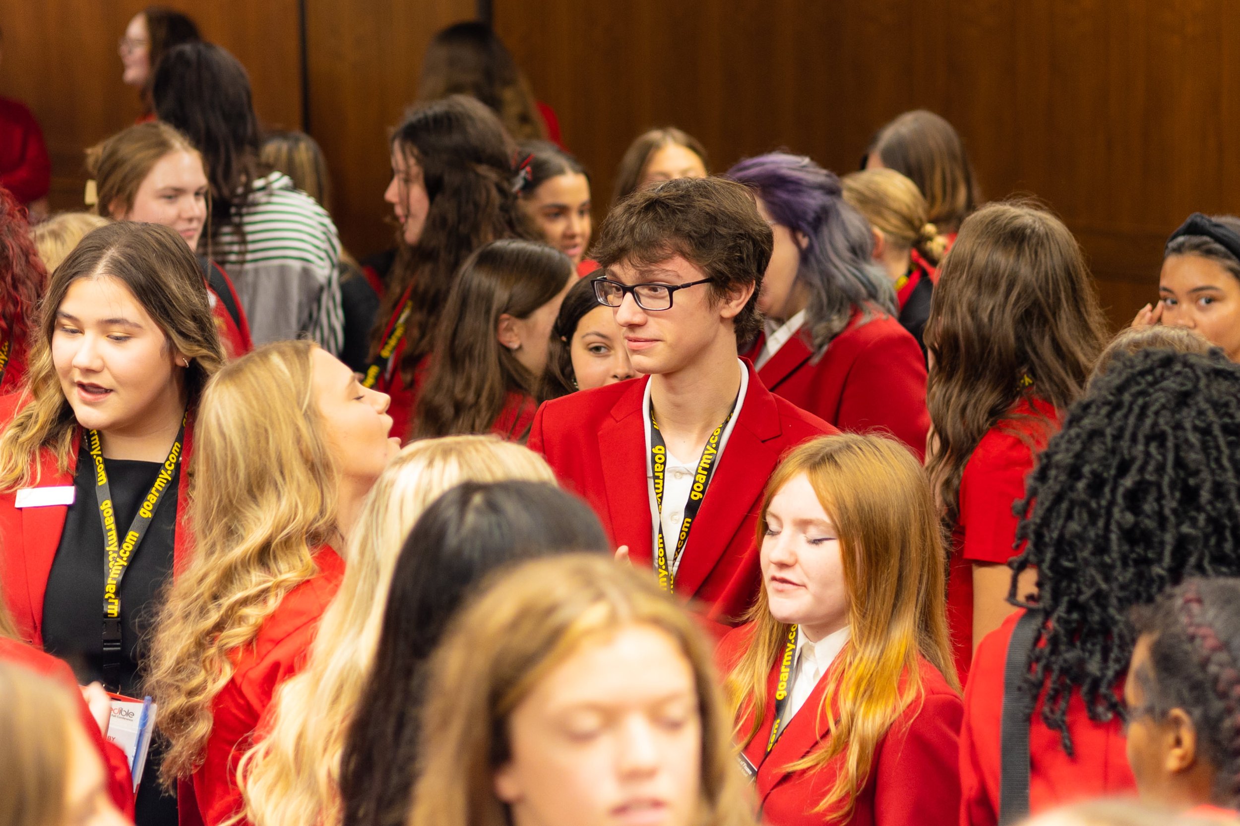 Group of young people wearing red blazers with yellow lanyards gathered at an indoor event.