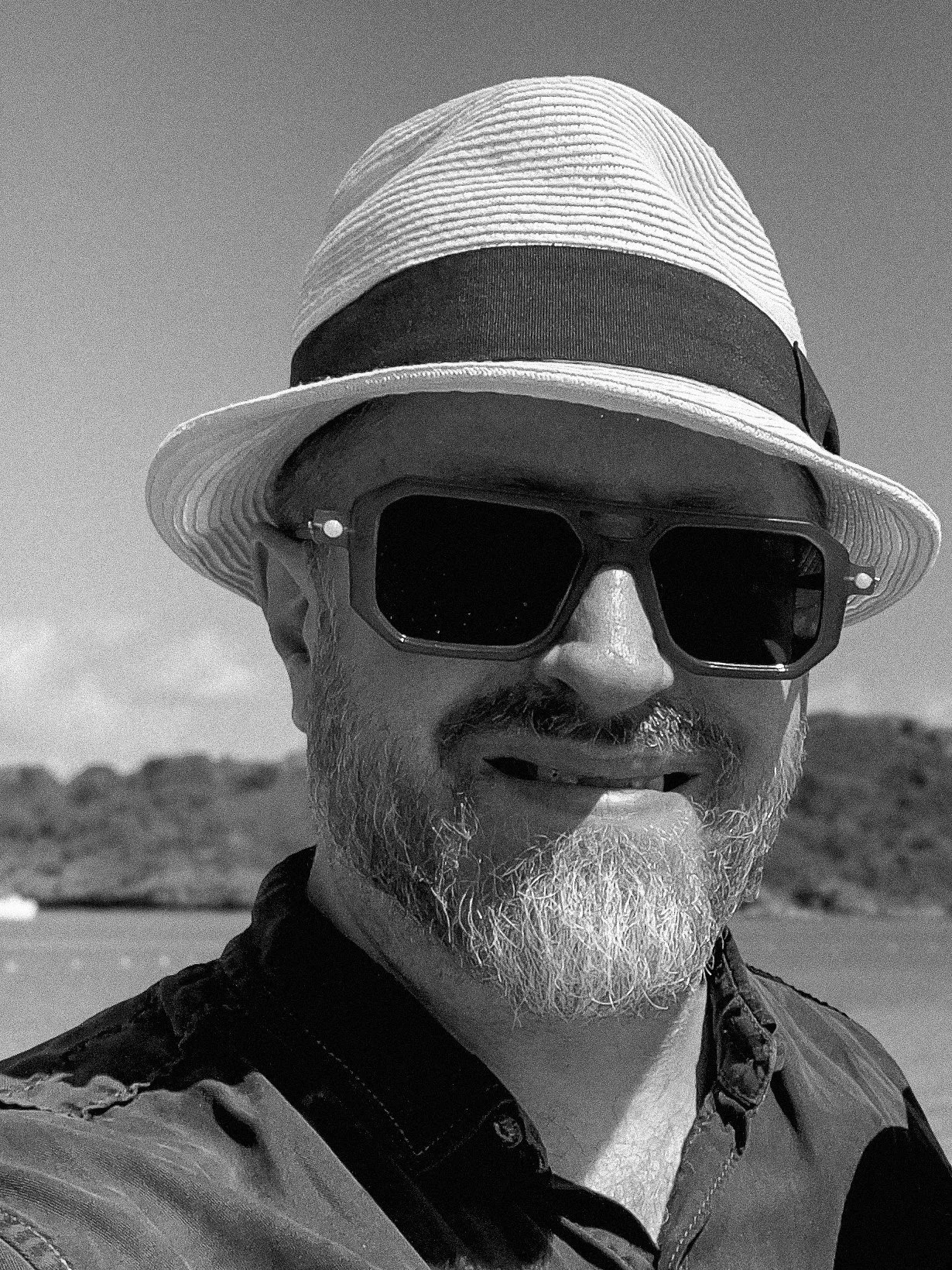 A man smiling on the beach wearing a straw hat, sunglasses, and a black jacket, with dunes and sky in the background.