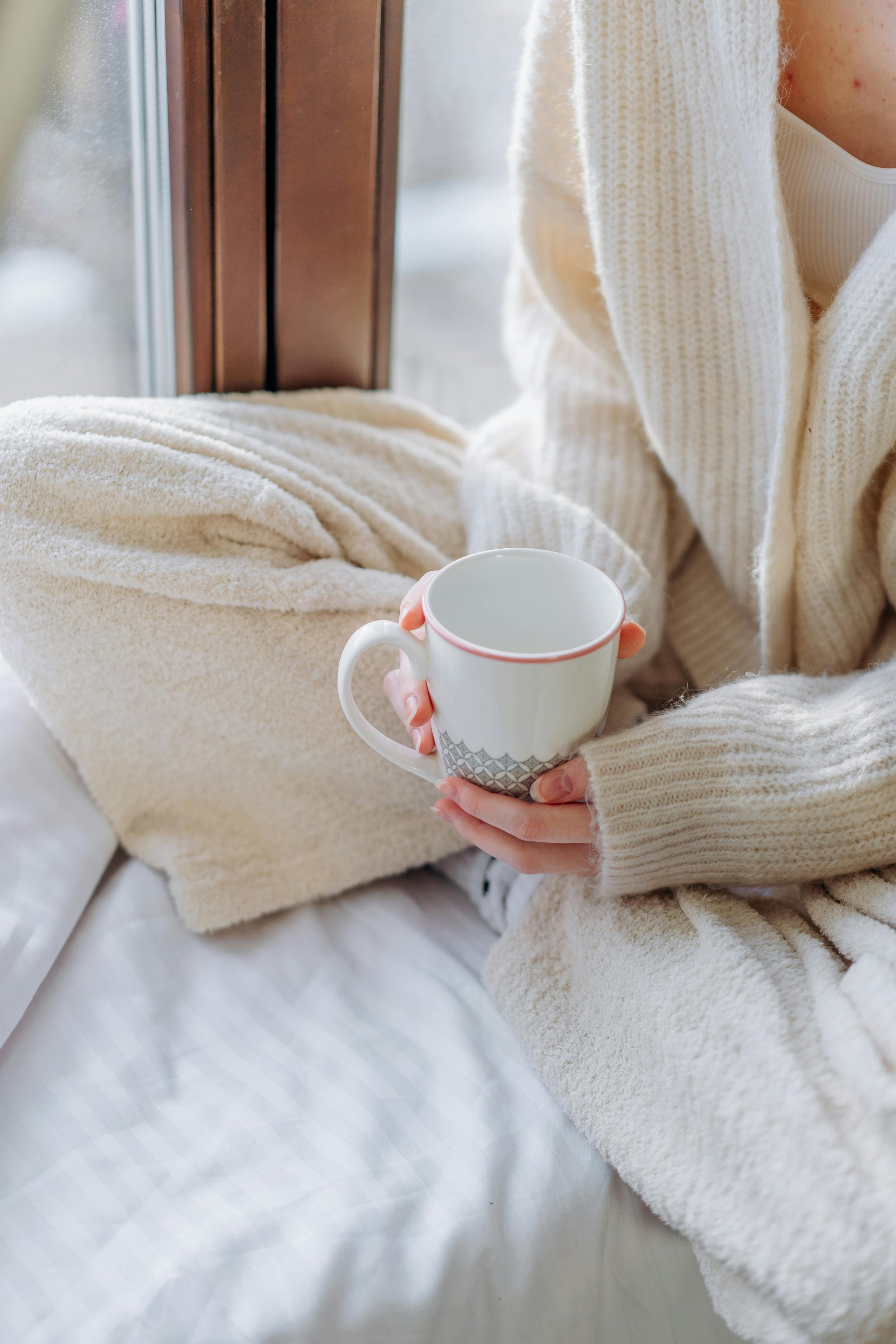Person in a cream sweater sitting by a window holding a mug in soft natural light