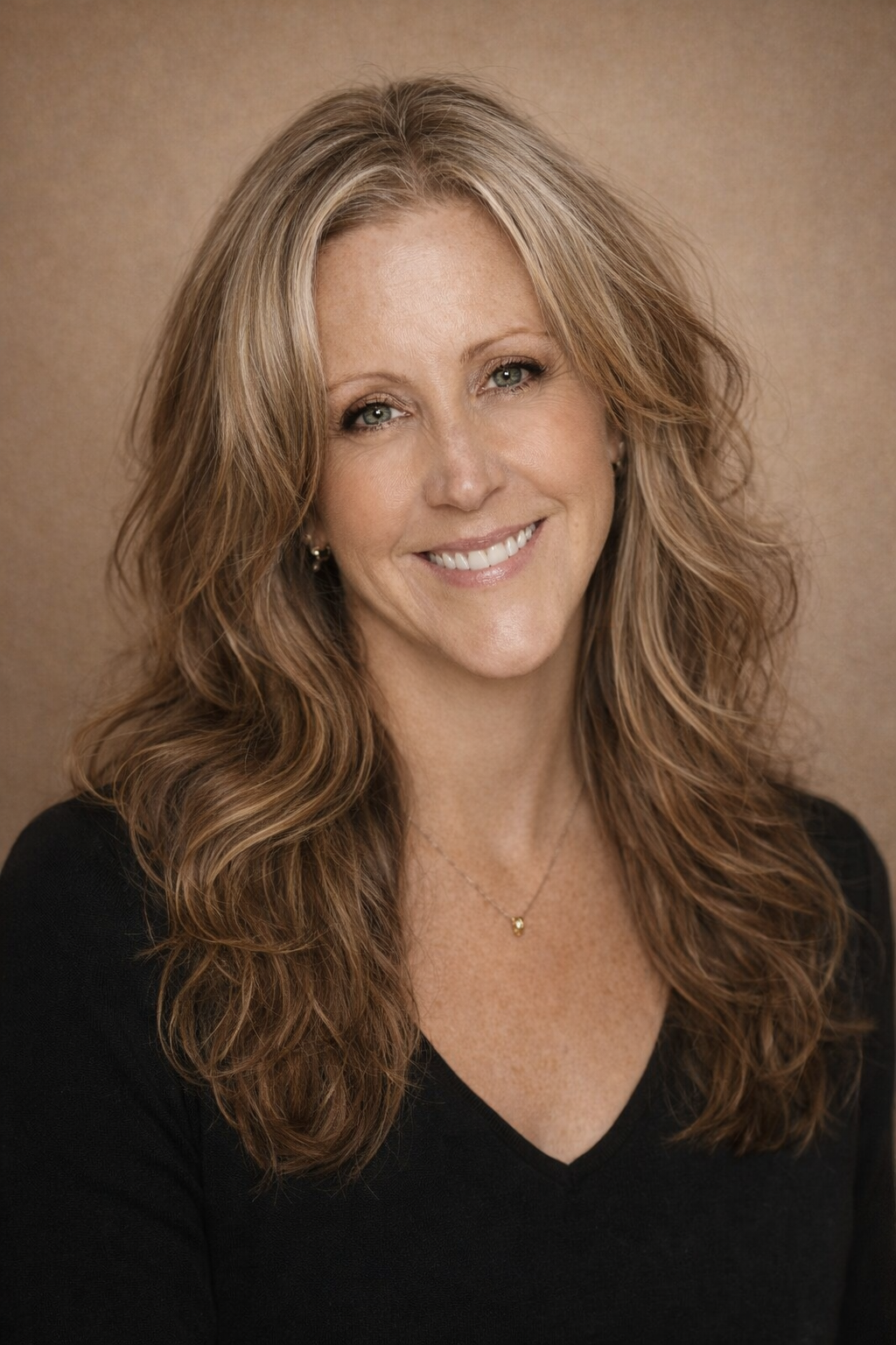 Headshot of a smiling woman with wavy, reddish-brown hair, wearing a black top and a small gold necklace, against a neutral background.
