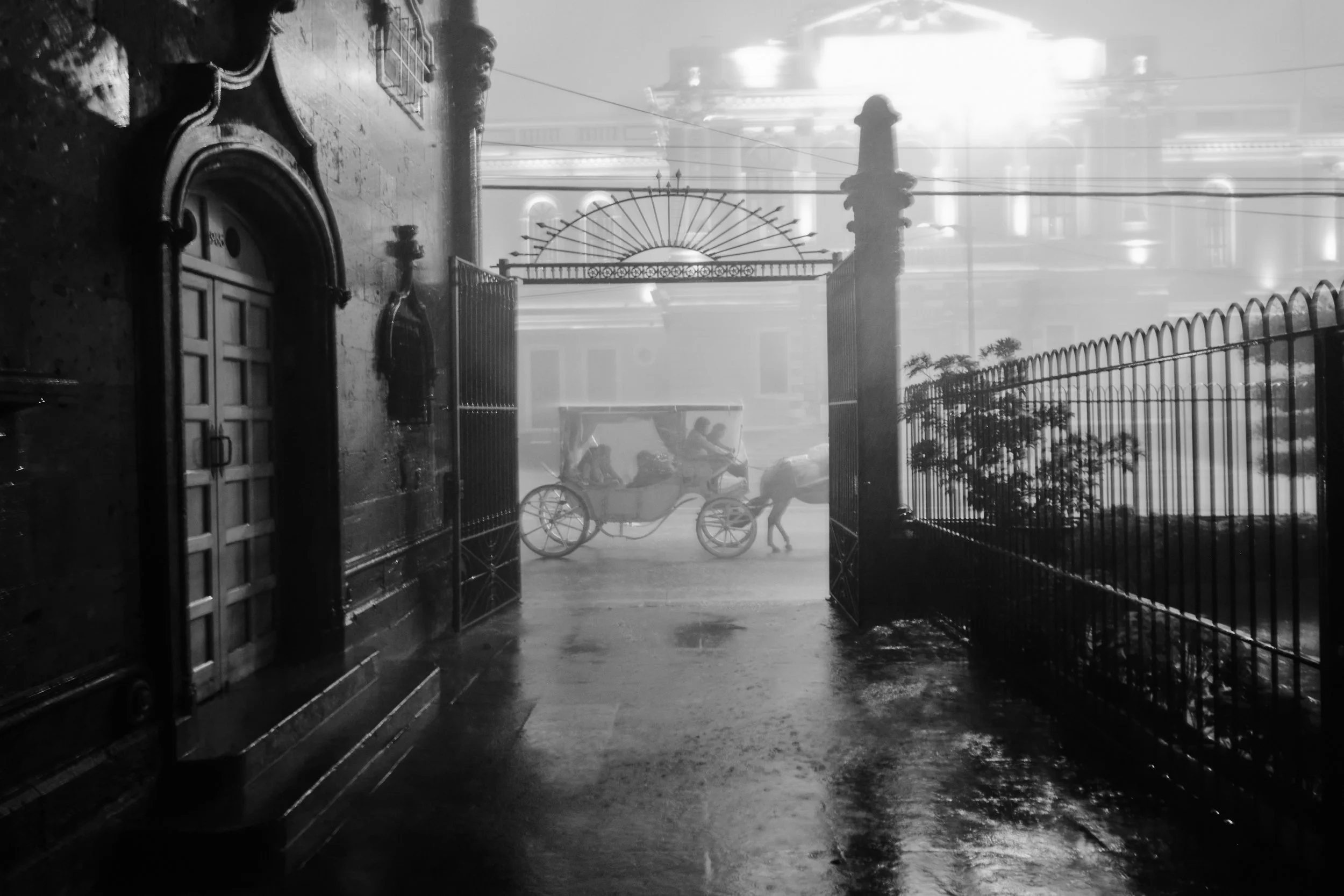 Escena en la calle con lluvia, vista desde una entrada con puertas y rejas, en la distancia un carruaje antiguo con caballos y un conductor, rodeado de un muro y arbustos, en ambiente oscuro y lluvioso. Expiatorio en Guadalajara, Jalisco, Mexico.