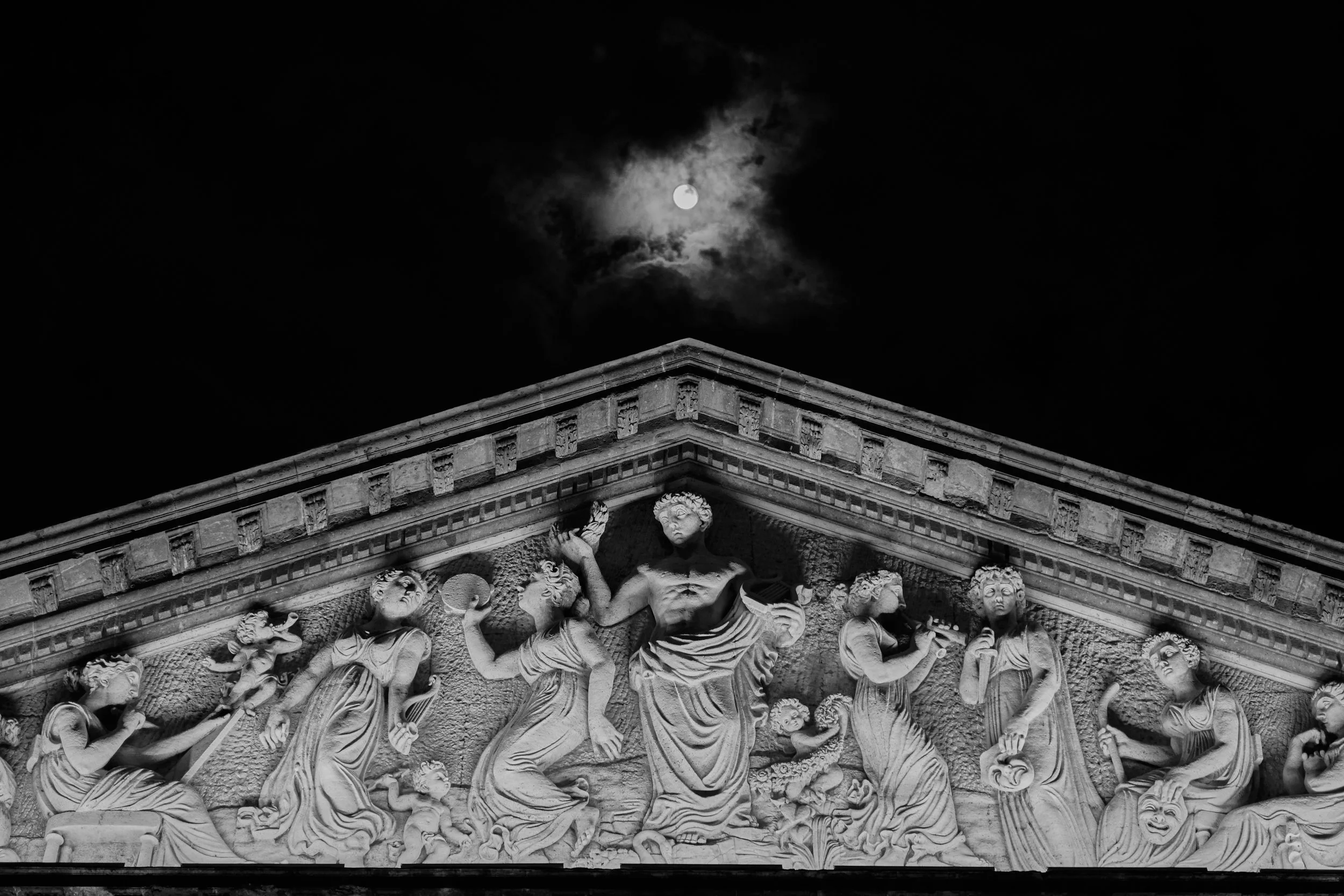 Detalle en la parte superior de un edificio antiguo con esculturas clásicas, bajo un cielo oscuro con luna llena y nubes. Teatro Degollado en Guadalajara, Jalisco, Mexico, con Luna Llena.