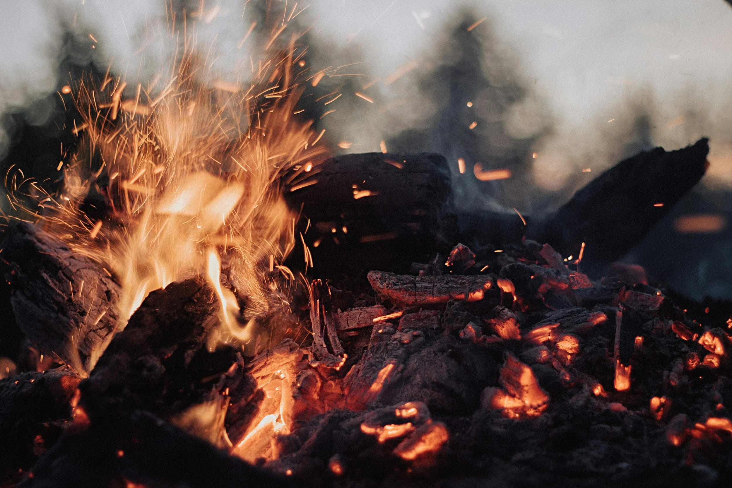 Close-up of a campfire with glowing embers, burning logs, and sparks flying into the air.