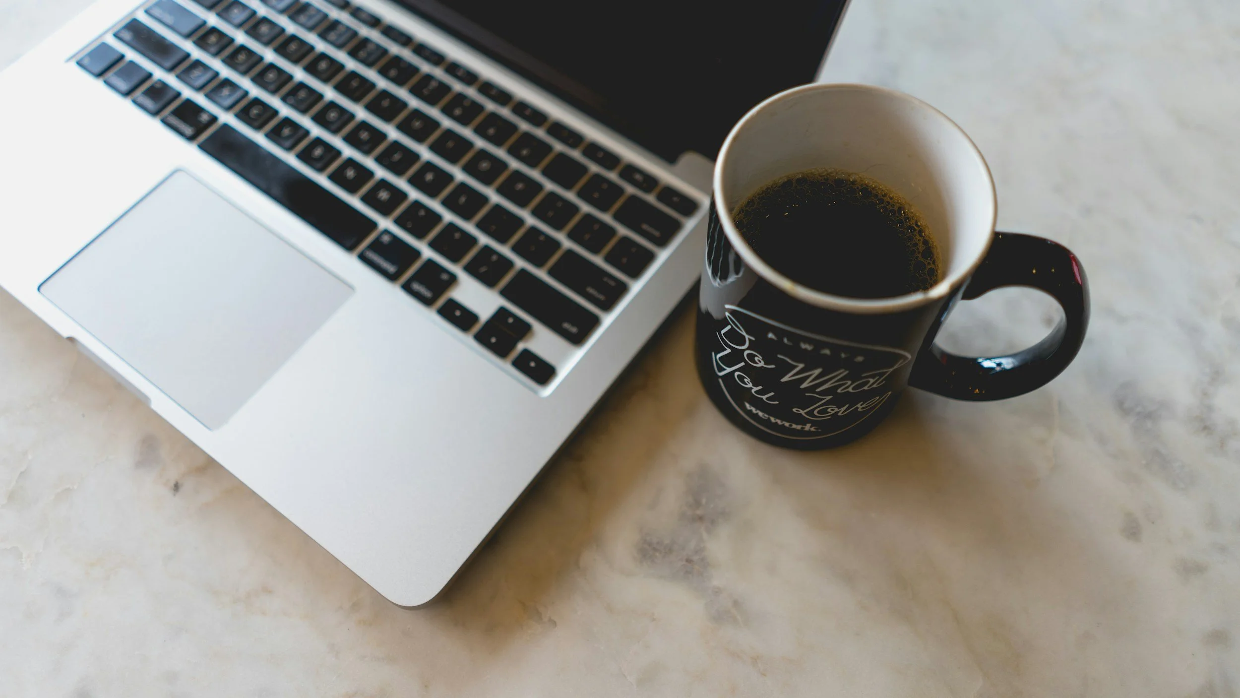 Open laptop with black keyboard and a white body next to a black coffee mug with text, placed on a beige marble surface.