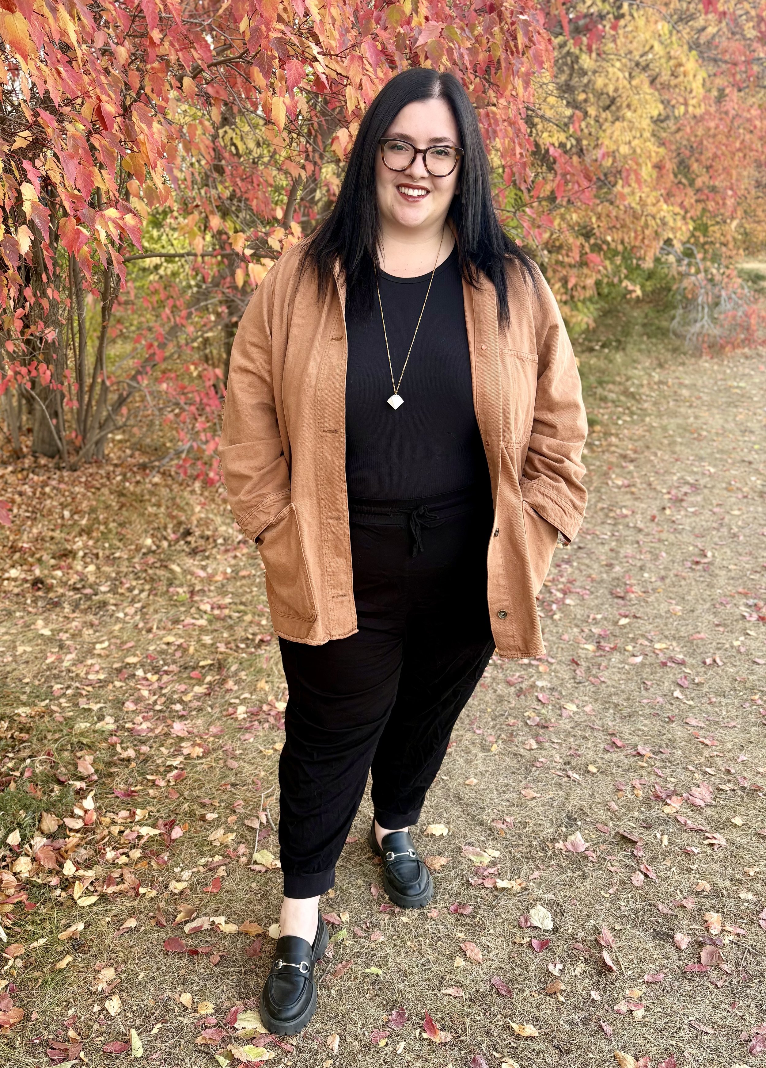 Therapist, Selena Arcovio, with black hair and glasses smiling outdoors in fall, wearing a black top, tan jacket, black pants, and black loafers, standing on a ground covered with fallen autumn leaves, with colorful fall trees in the background.