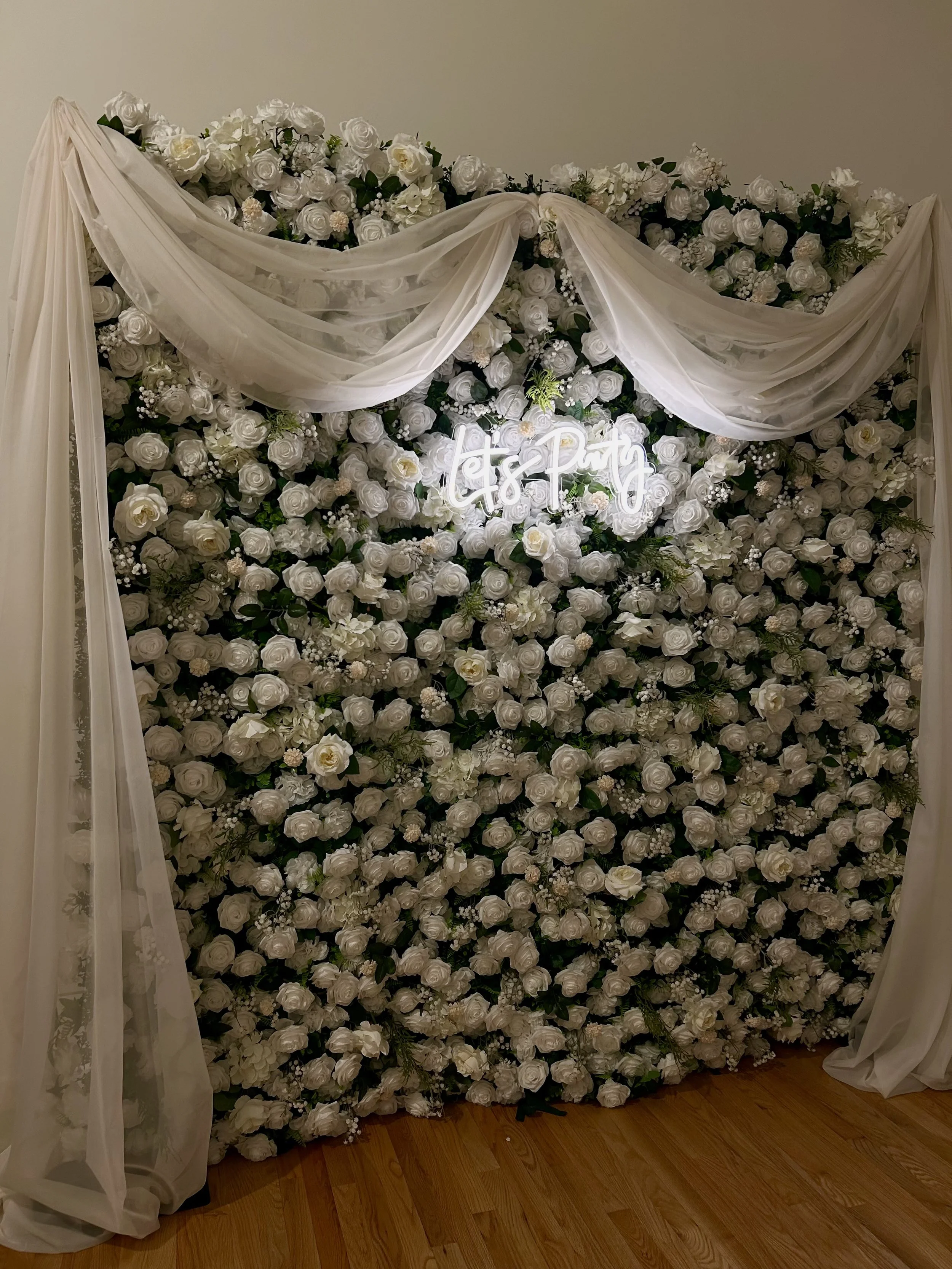 Flower wall with white roses, greenery, sheer drapes, and a neon sign that says 'let's party'.