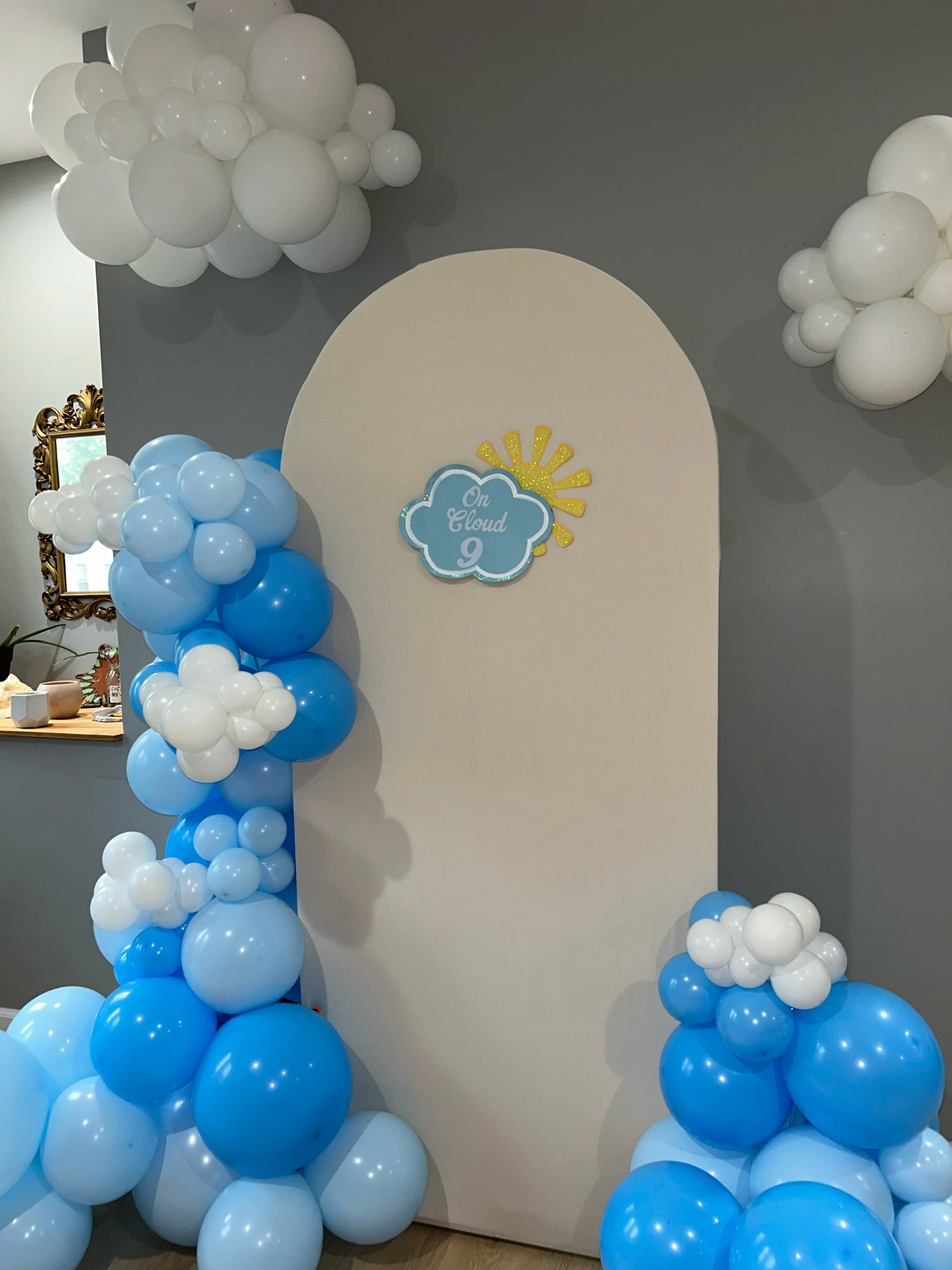 Blue and white balloon arch and clusters with a white backdrop and cloud-themed sign reading 'On Cloud 9' for a celebration.