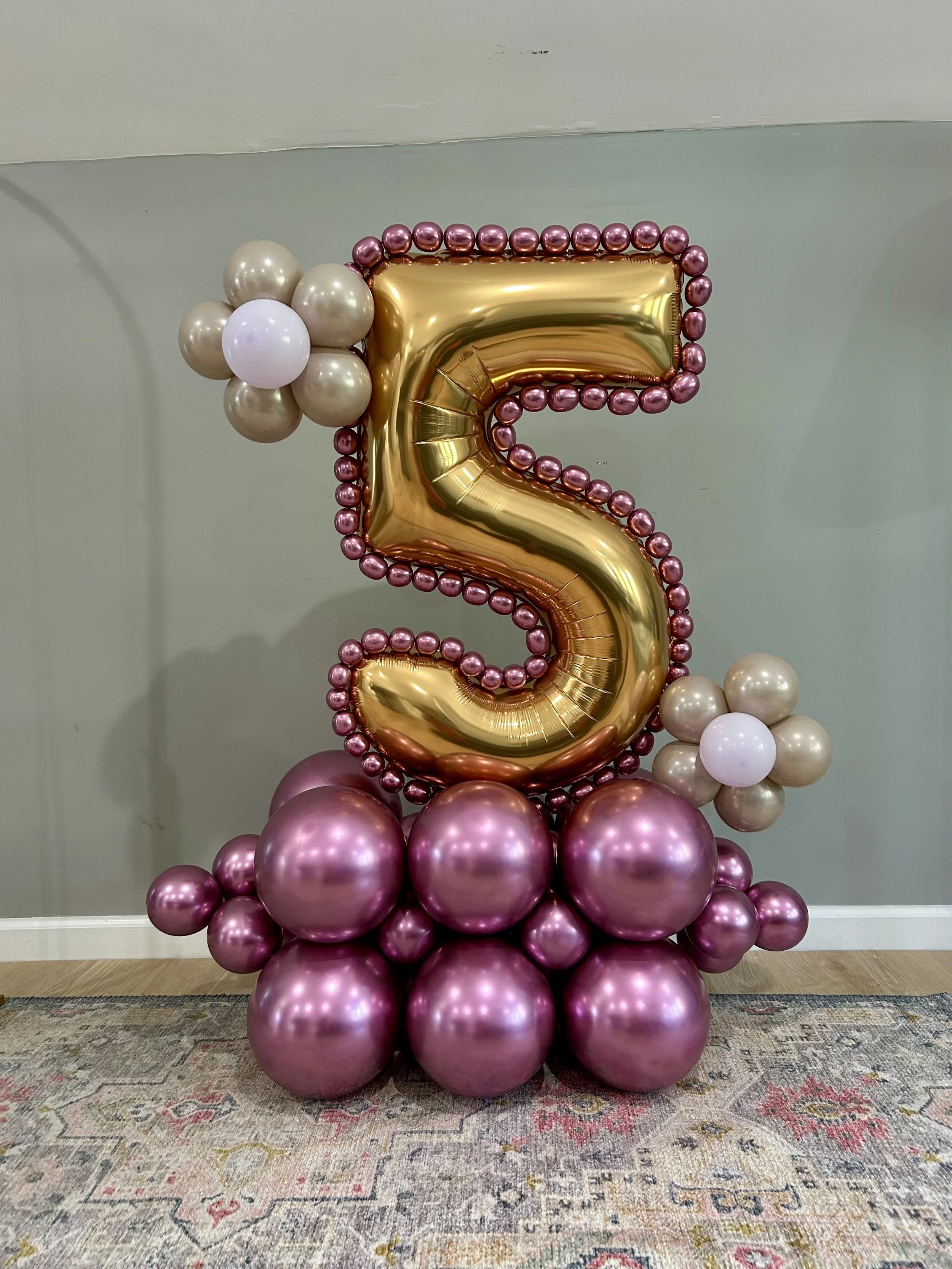 A large gold foil balloon shaped number five, surrounded by pink and beige balloons, with pink balloons forming a base and beige balloons on the sides, arranged for a celebration.
