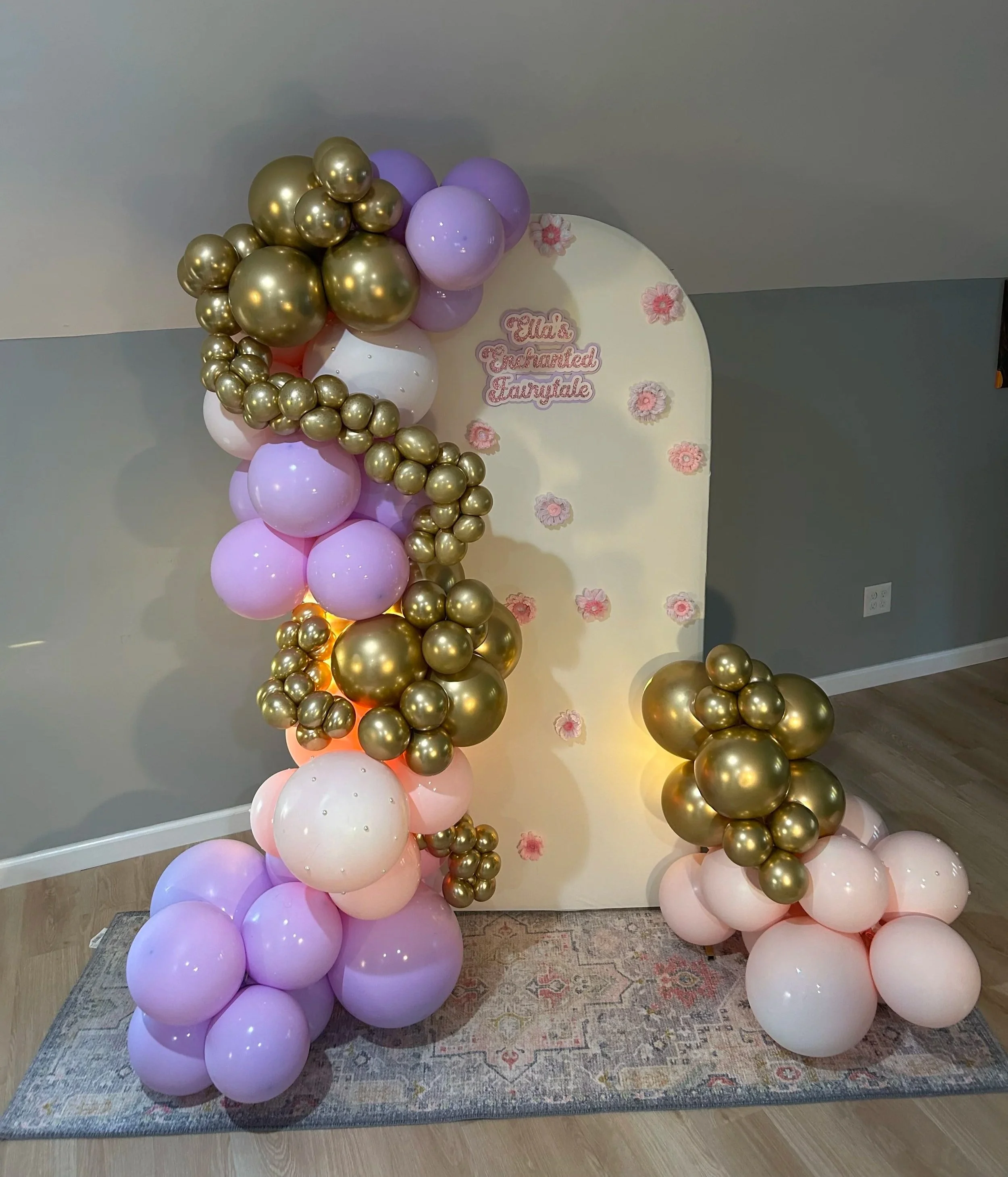 Balloon display with pink, white, purple, and gold balloons created for a celebration, with a backdrop that has pink flower decorations and a sign reading 'Ella's Chachareted Earsdale'.