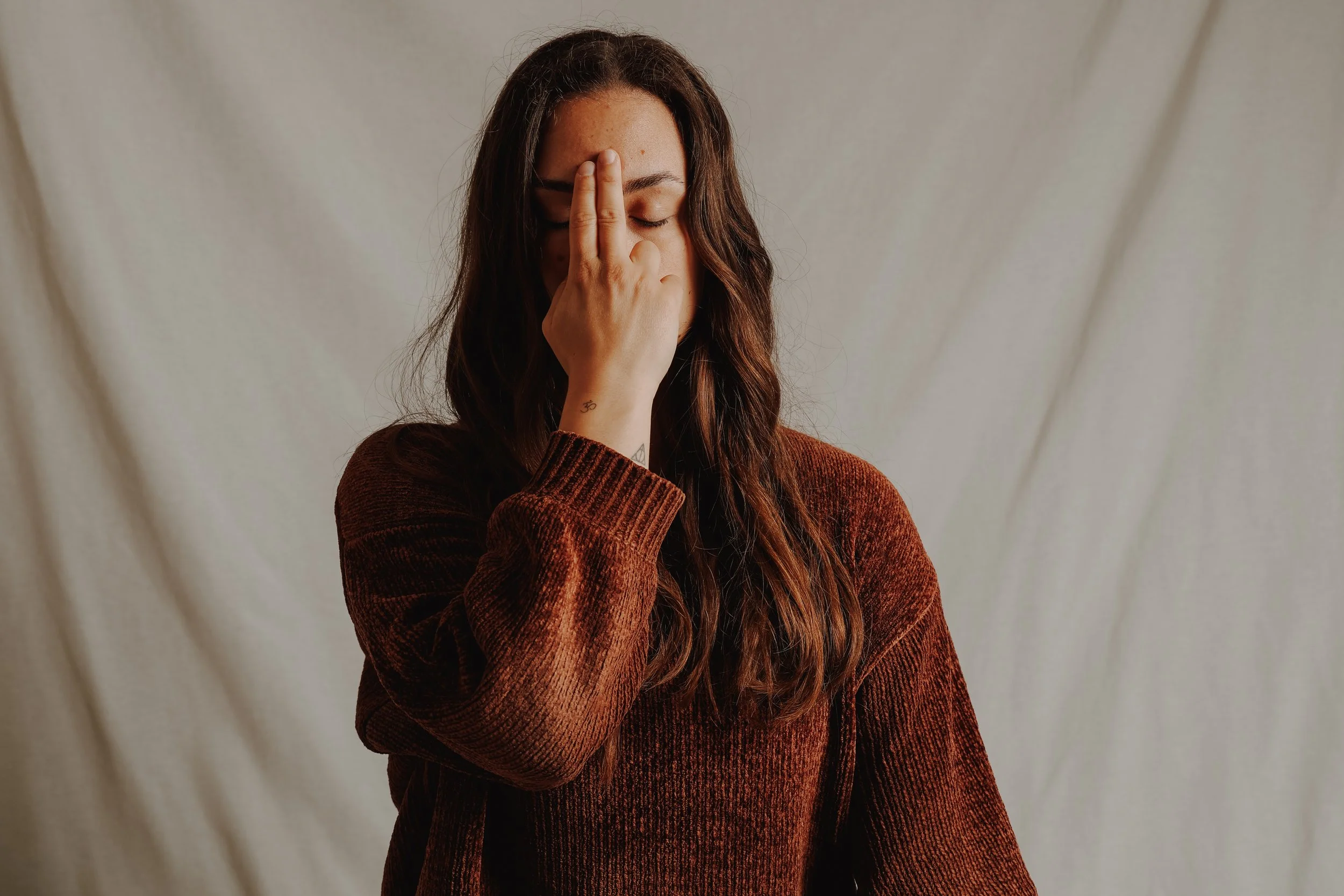Junge Frau mit braunen Haaren trägt einen braunen Pullover, hält die Hand vor das Gesicht und vermeidet Blickkontakt.