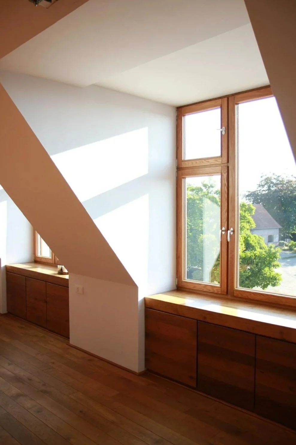 Ein Zimmer mit großen Fenstern, Holzboden und eingebauten Holzschränken unter den Fenstern, helle Wände und viel natürliches Licht, das durch die Fenster strömt.