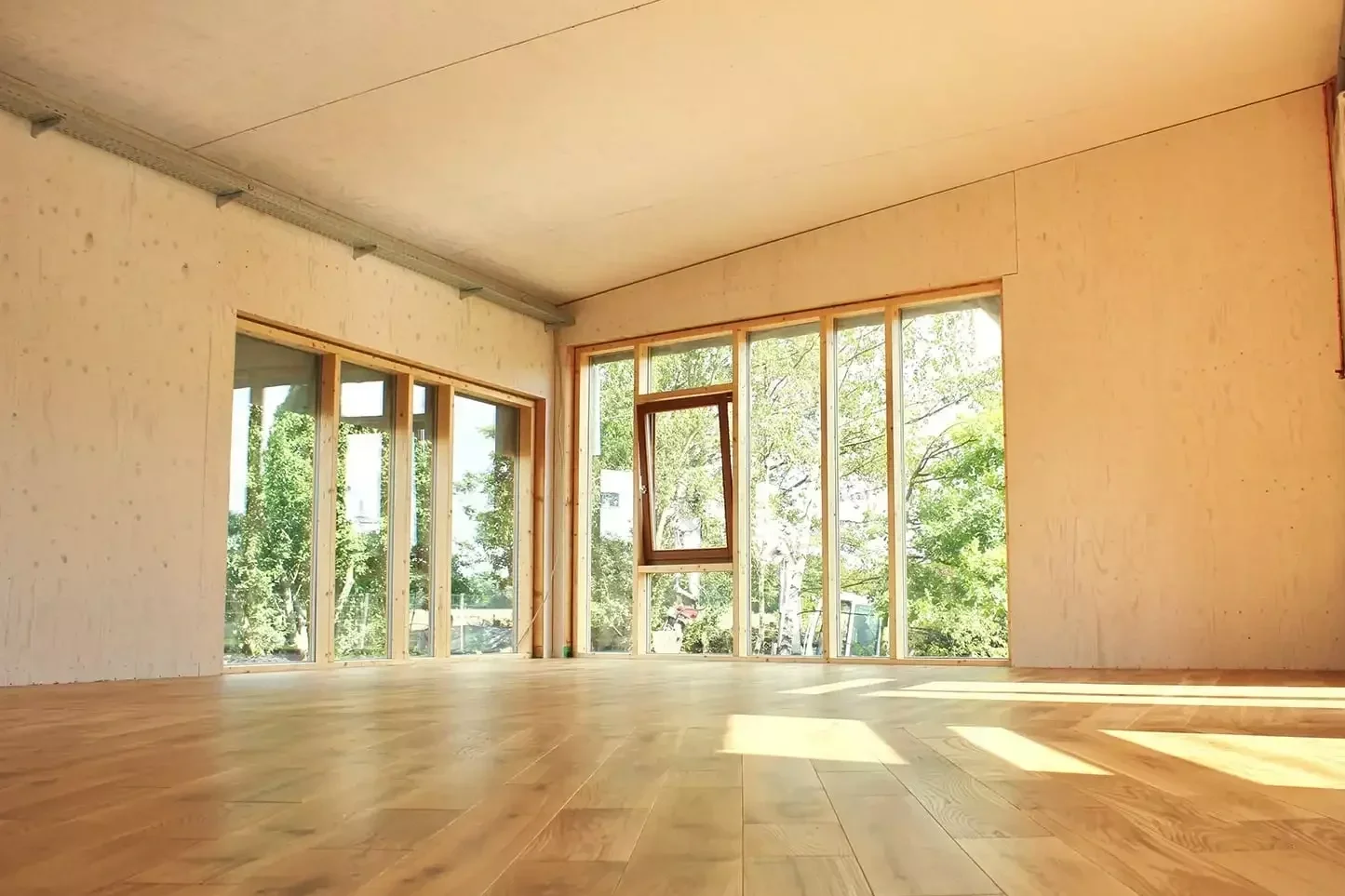 Unvollendeter Raum mit großen Fenstern und Holzfußboden, Blick nach draußen auf Bäume.