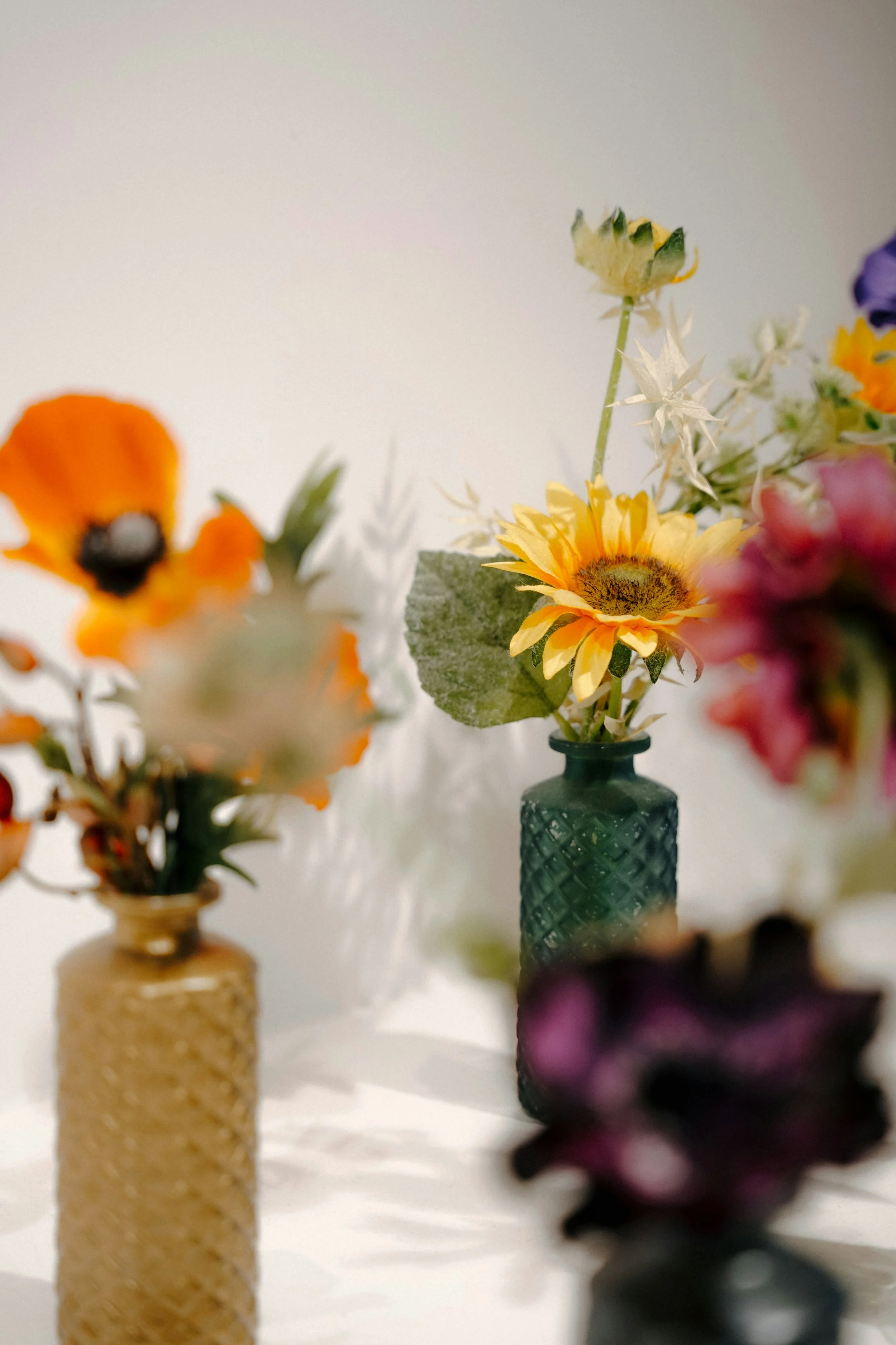 Verschiedene Blumen in bunten Vasen auf einem weißen Tisch vor einem hellen Hintergrund.