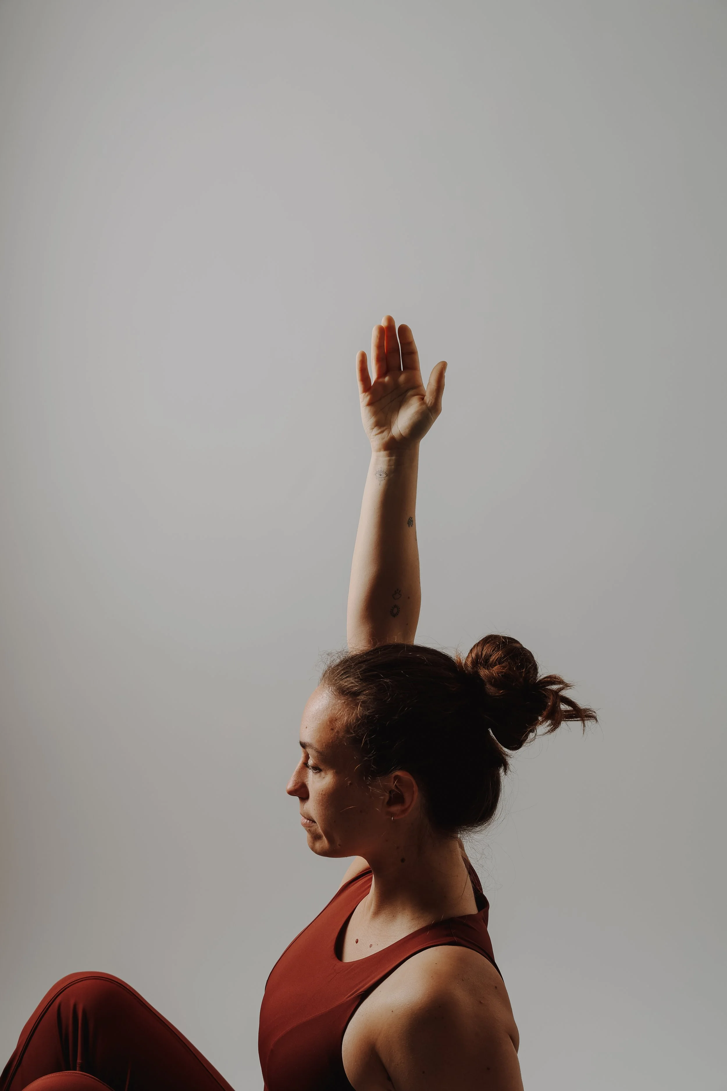 Frau in Yoga-Positur mit erhobener Hand, trägt rote Sportkleidung, gegen grauen Hintergrund.