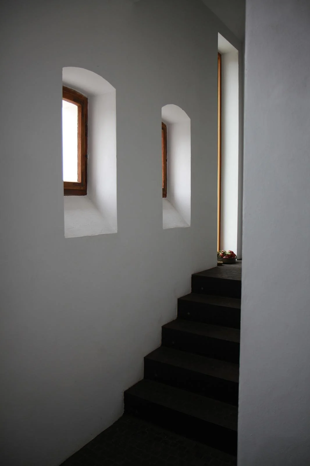 Schwarze Treppen vor weißen Wänden mit drei kleinen rechteckigen Fenster.