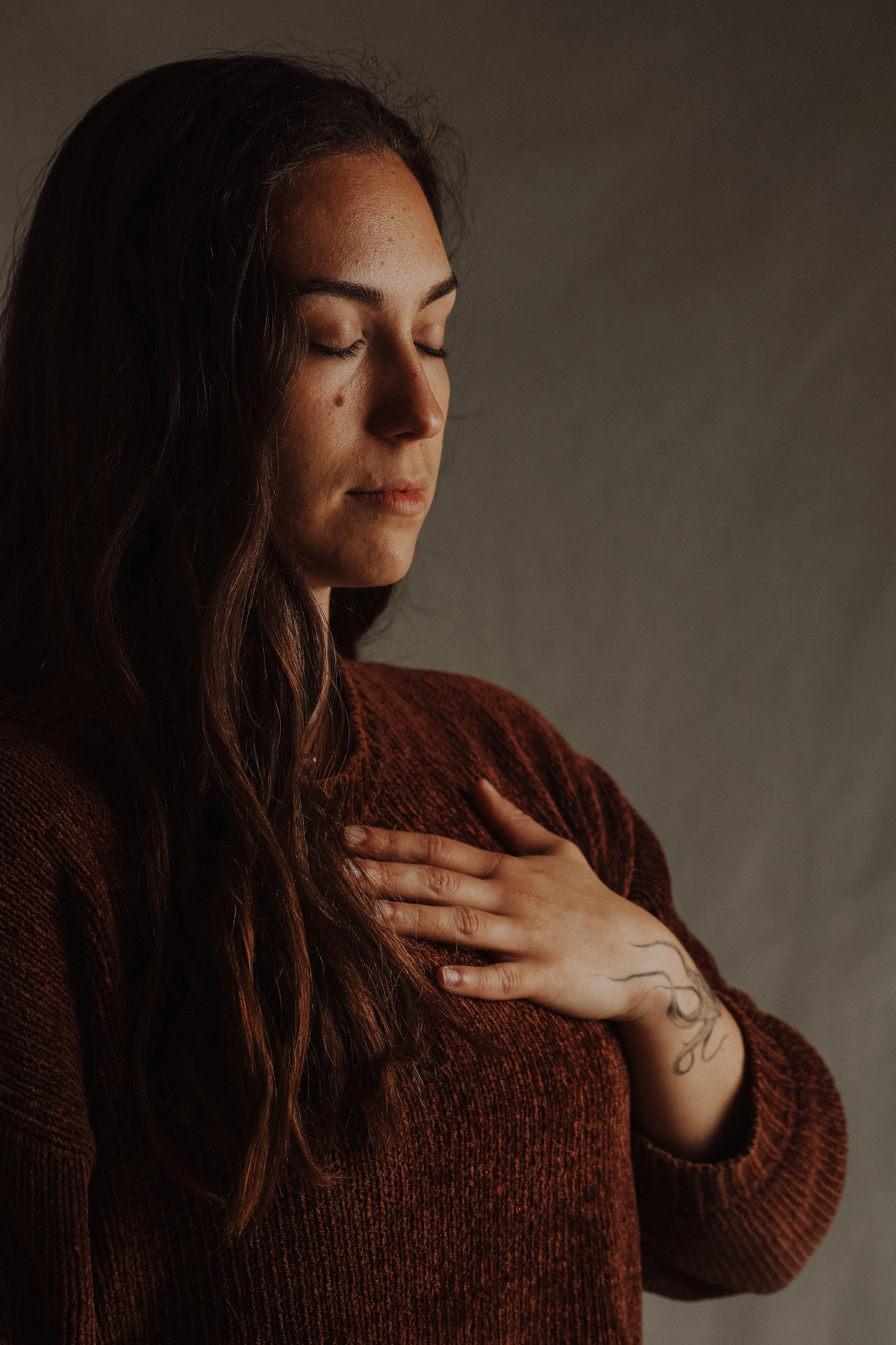 Ein junge Frau mit geschlossenen Augen und Hand auf der Brust, trägt einen braunen Pullover, hat lange braune Haare und eine Tätowierung am Handgelenk, vor grau hintergrund