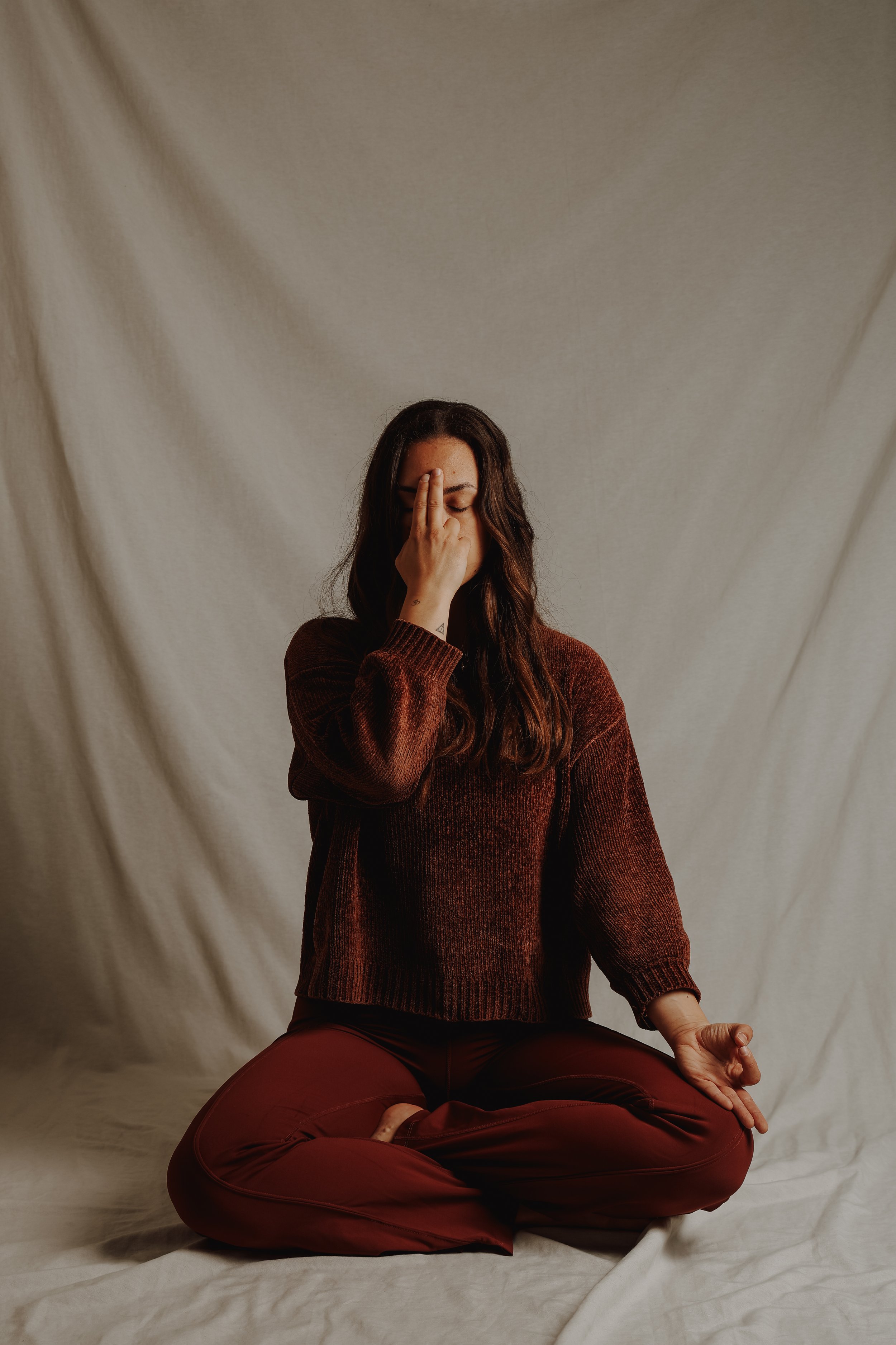 Frau sitzt im Schneidersitz, trägt braune Kleidung, hat die Augen geschlossen, die Hand vor das Gesicht gehalten, vor neutralem Hintergrund.