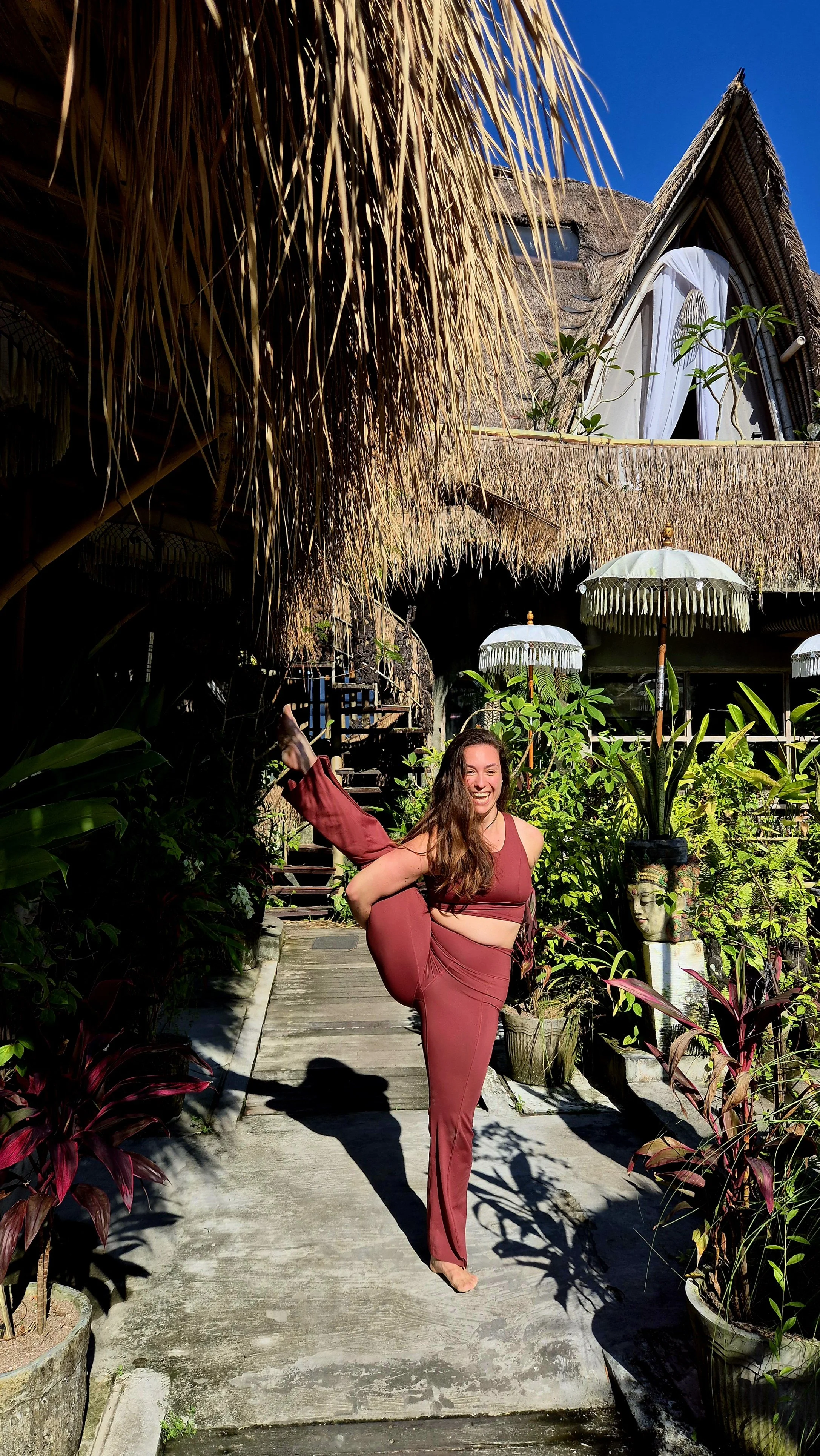 Eine lachende Frau in einem roten sportlichen Outfit macht einen Handstand auf einem Weg in einem tropischen Garten mit Pflanzen, Sonnenschirmen und einem Holzhaus im Hintergrund.