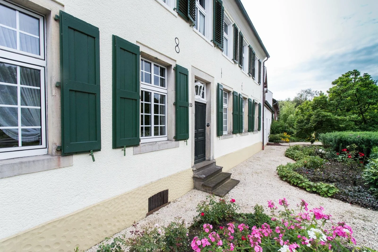 Ein Haus mit weißen Wänden, grünen Fensterläden und einem schwarzen Eingangstür, umgeben von einem Garten mit bunten Blumen und Bäumen.