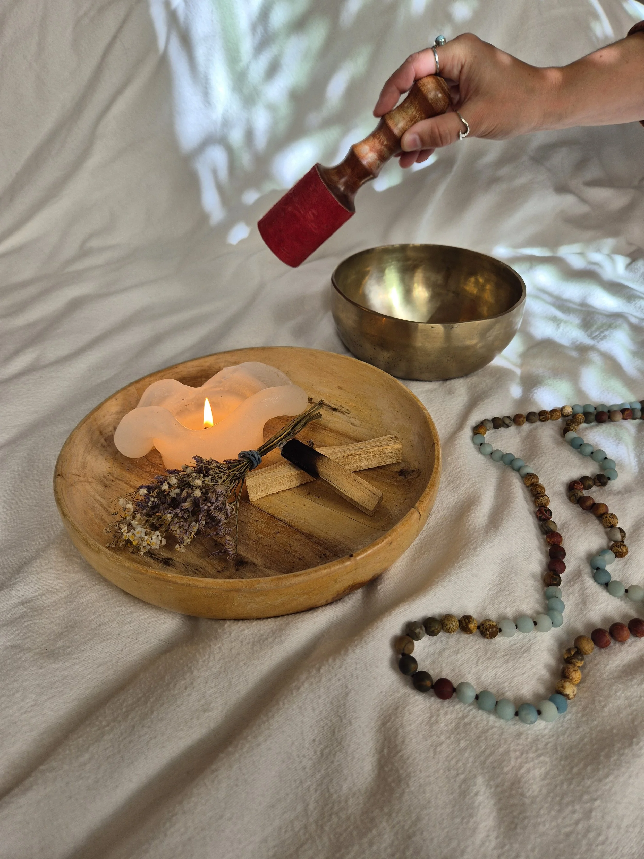 Hand hält einen Polsterklopfer über eine goldene Klangschale, daneben ein Holztablett mit Kerze, Lavendel und Holzklötzen, eine Bead-Kette liegt auf weißem Stoff.