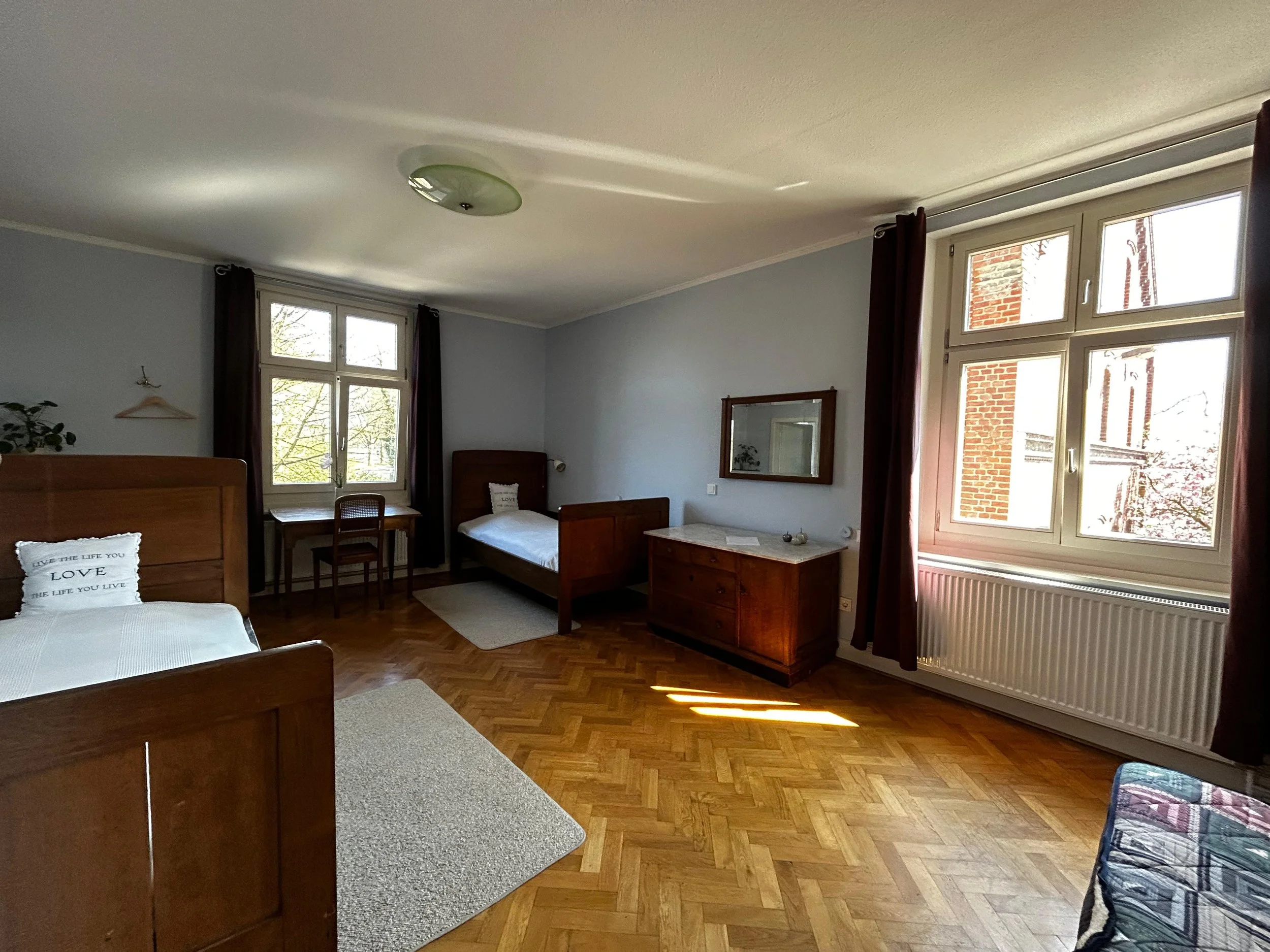 Ein hell erleuchteter Schlafraum mit zwei Einzelbetten, einem Schreibtisch vor einem Fenster, einem Kleiderschrank, einem Sideboard mit Spiegel und einem großen Fenster mit dunklen Vorhängen.