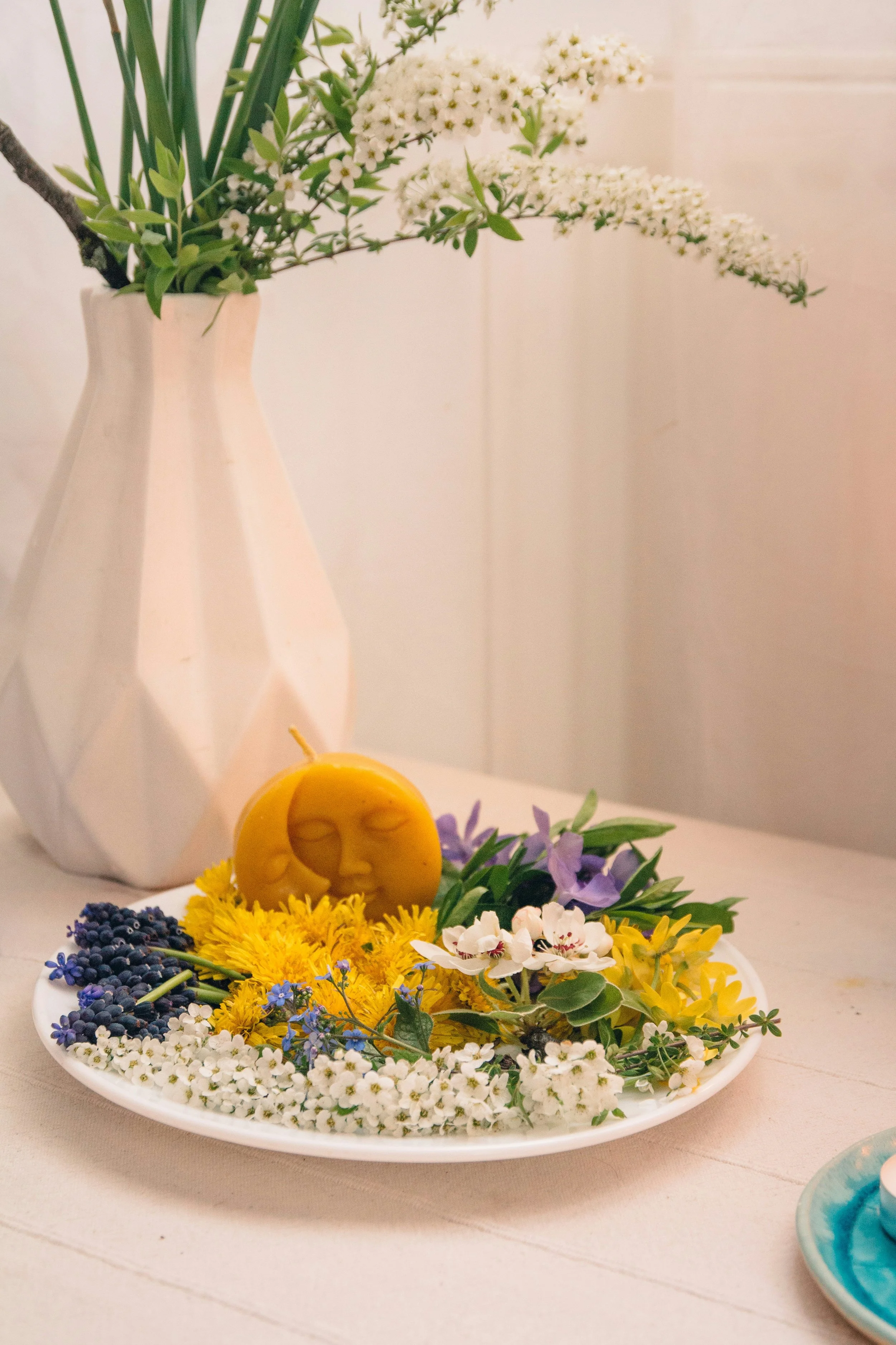 Vase mit weißen Blumen und grünen Blättern im Hintergrund, auf einem weißen Tisch liegt ein weißer Teller mit verschiedenen bunten Blumen und einer gelben Kerze, die wie eine lachende Sonne gestaltet ist.