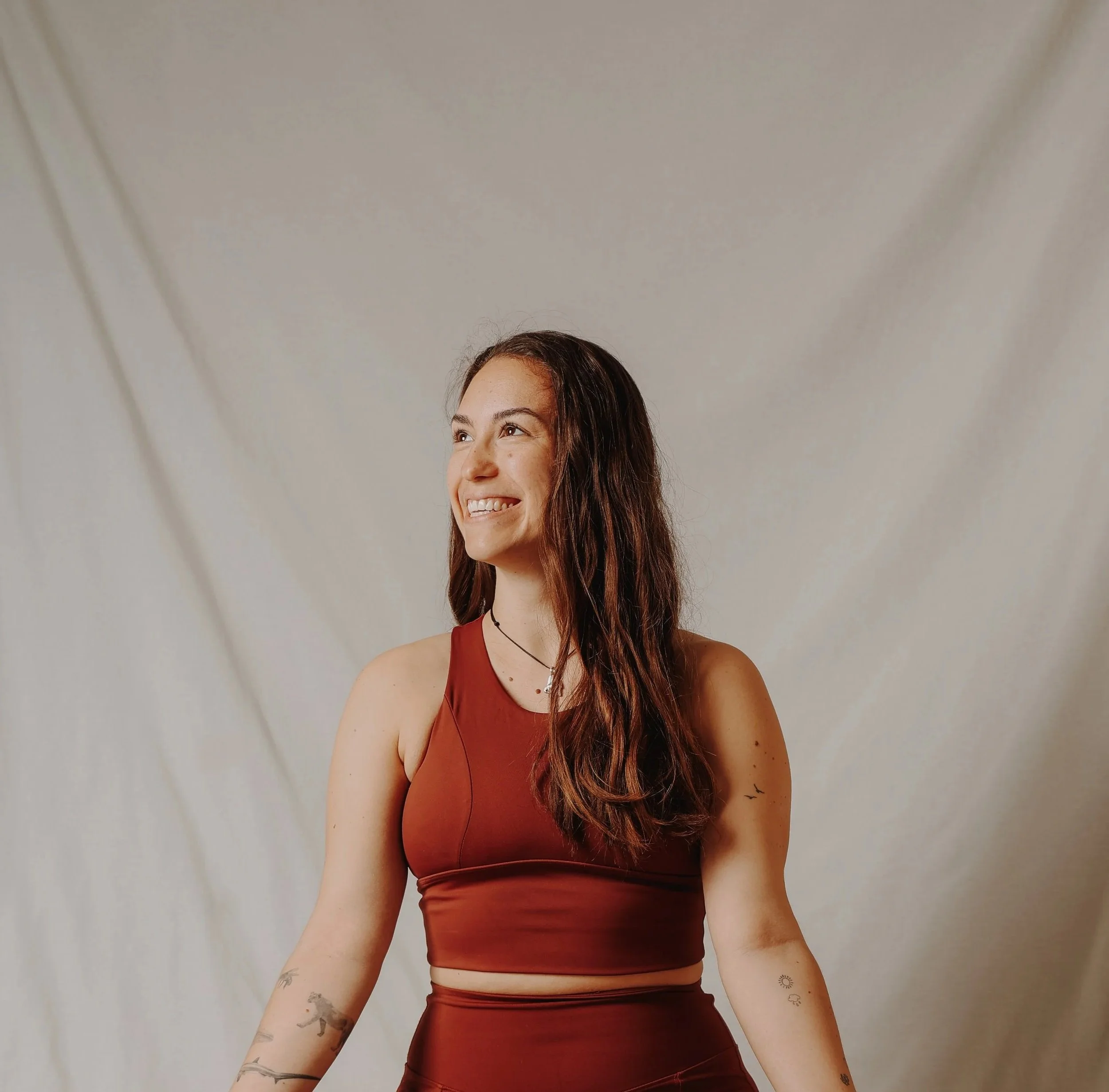 Eine junge Frau mit langen braunen Haaren, die in einem rot-braunen Sportoutfit lächelt, vor hellem Hintergrund.