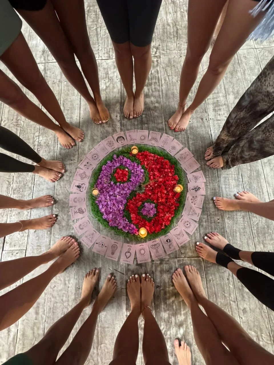 Menschen stehen im Kreis um ein Mandala aus Blumen, Kerzen und Karten auf dem Boden.